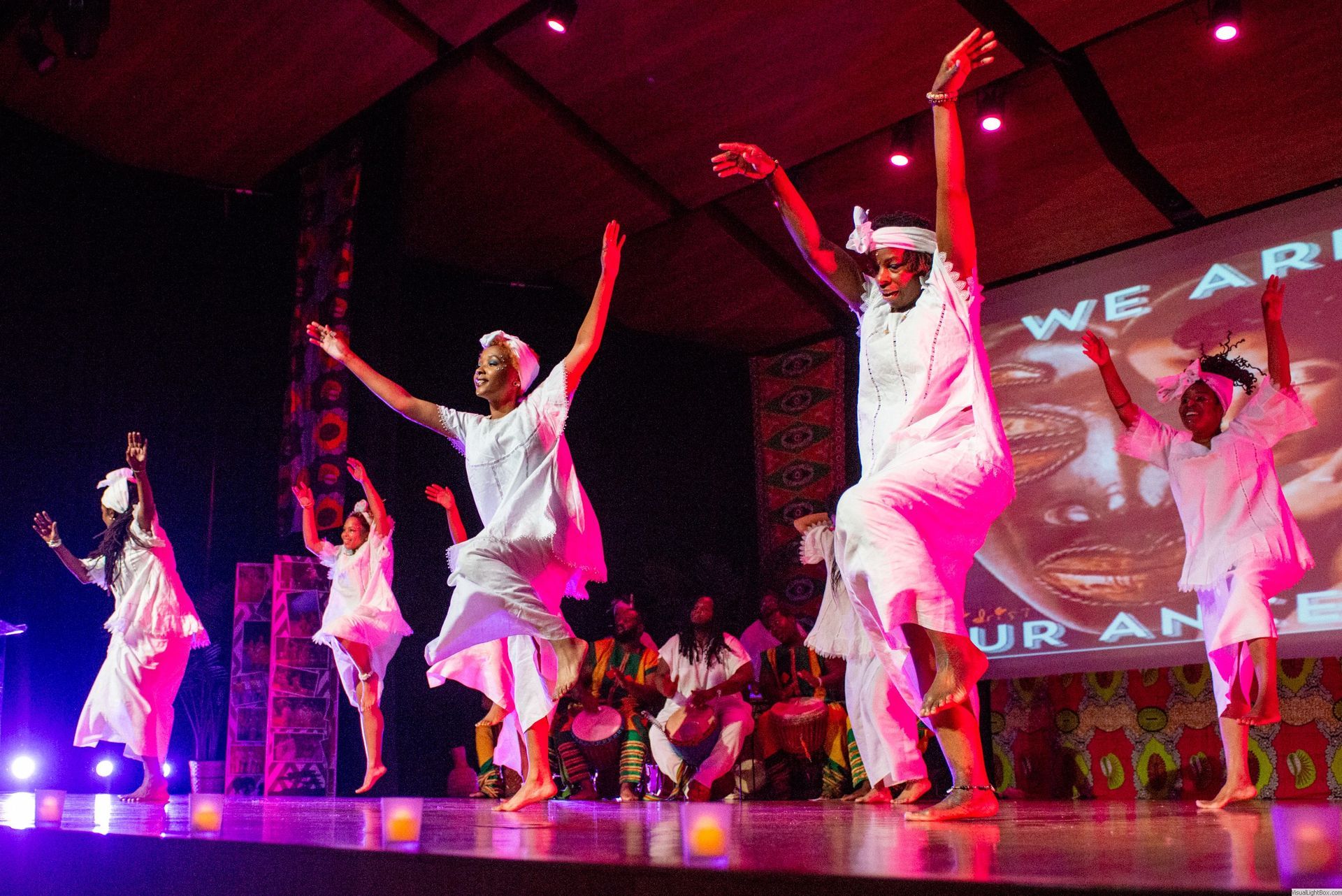 Dancers in white costumes perform on stage with arms raised; a screen in the background reads 