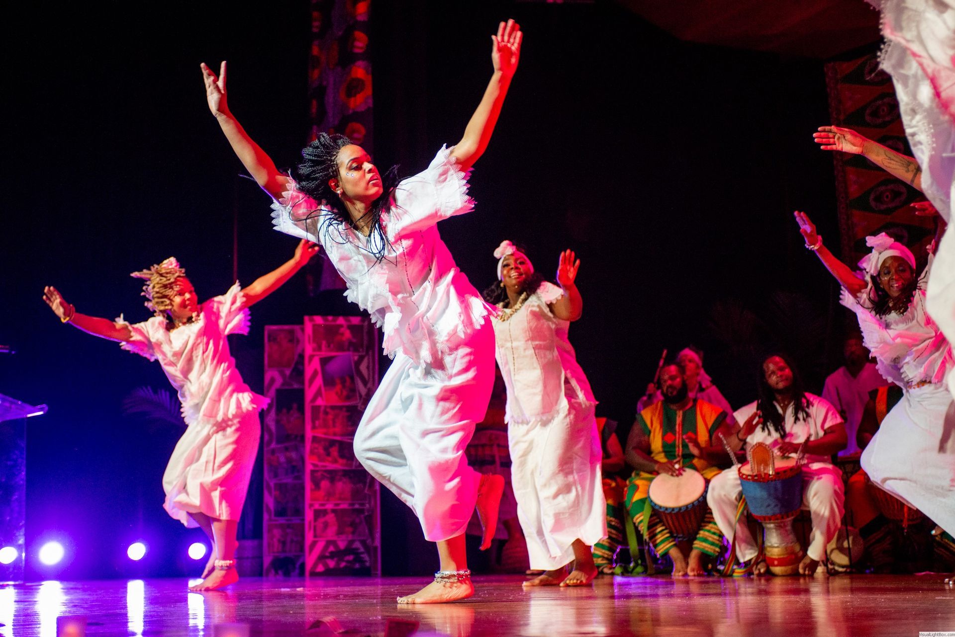 Dancers in white outfits with arms raised perform on a stage. Musicians play drums in the background.