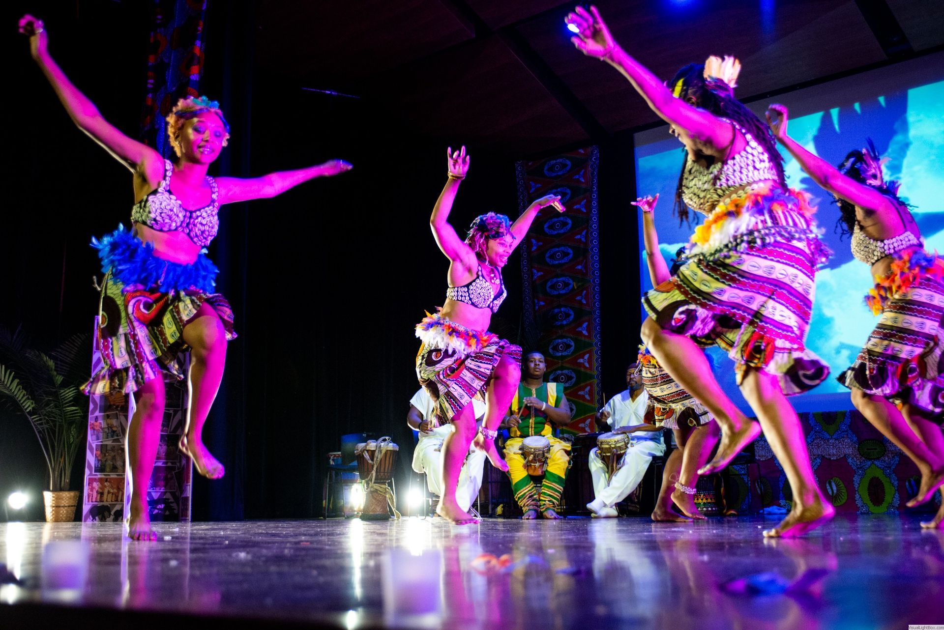 African dancers in colorful outfits leap on stage under bright lights.