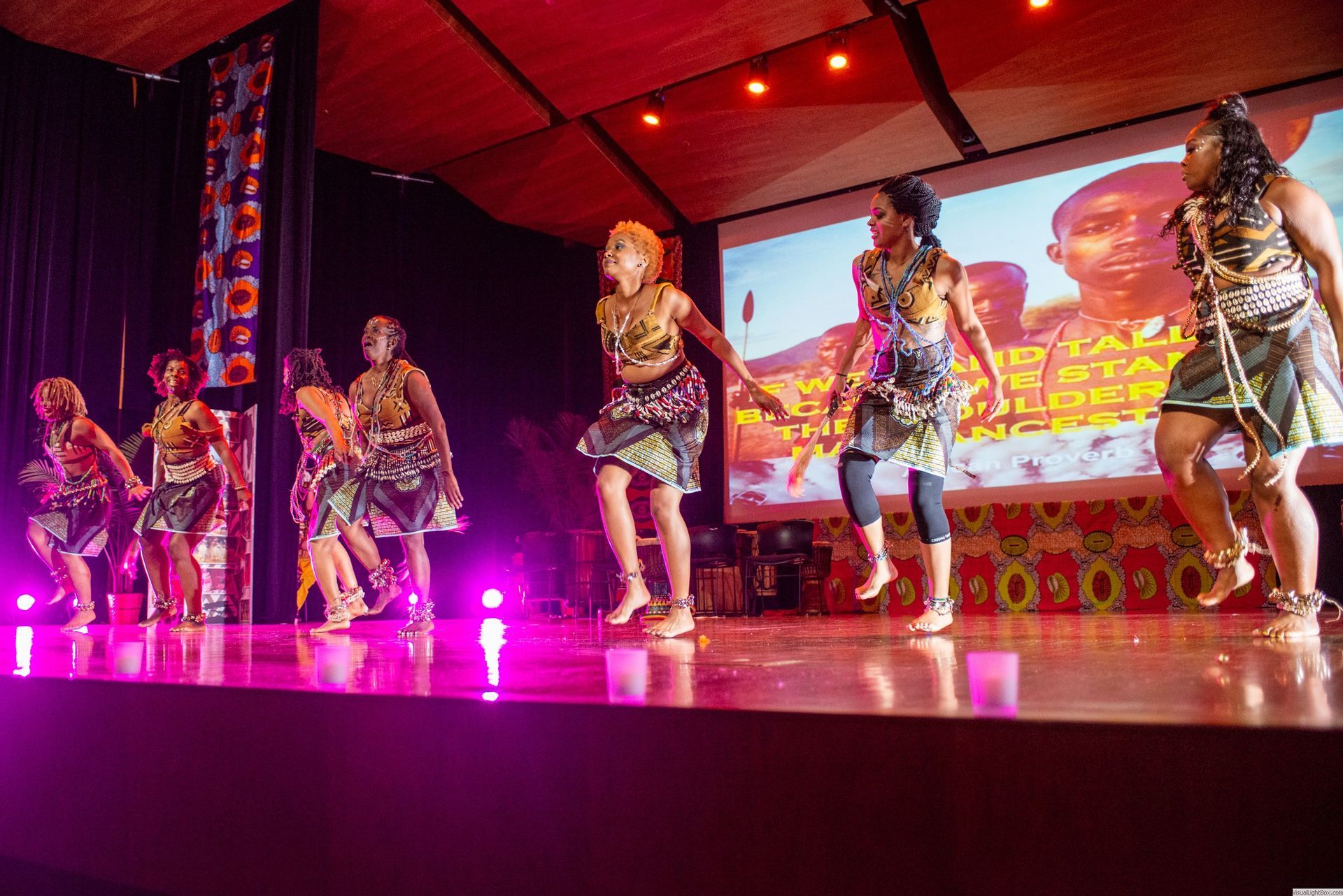 Dancers on a stage in colorful outfits, performing with a projection backdrop. Lit with pink and purple lights.