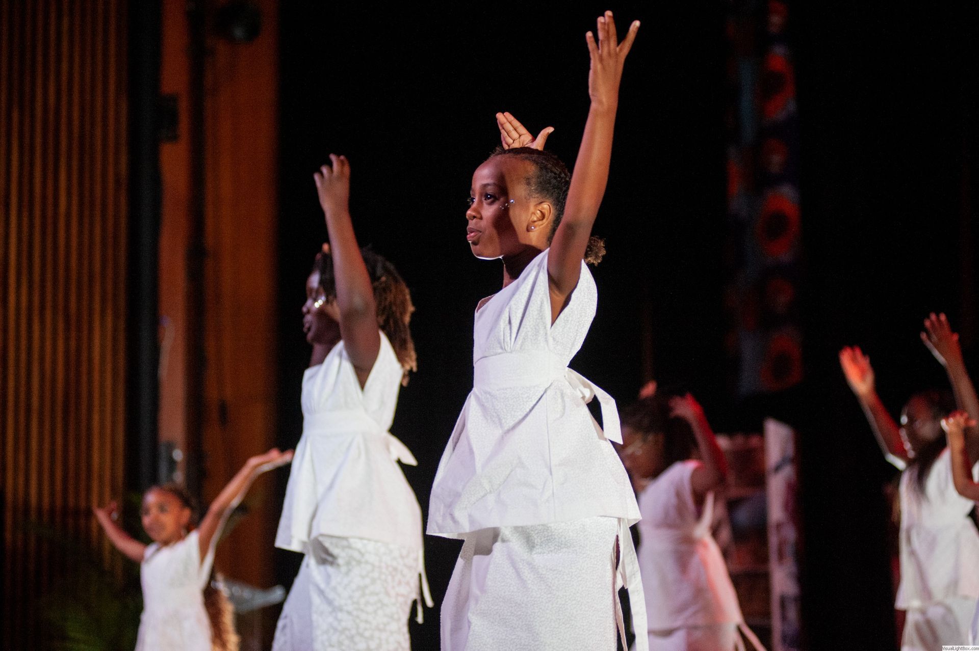 Young dancers in white dresses performing on stage, arms raised.