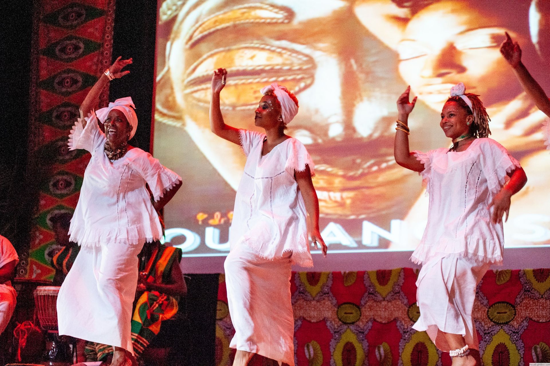 Three women in white performing a dance on stage with an African-themed backdrop.