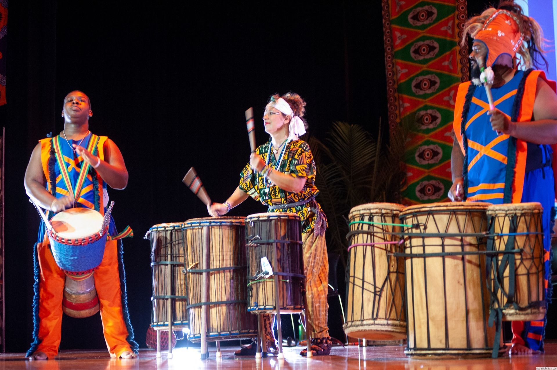 Three performers in colorful African-style outfits playing drums onstage.