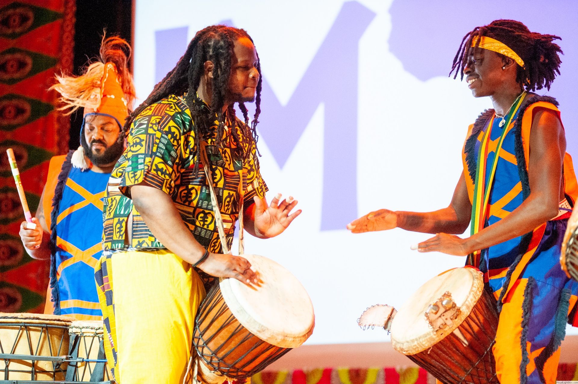 Three African drummers perform on stage, wearing colorful traditional clothing.