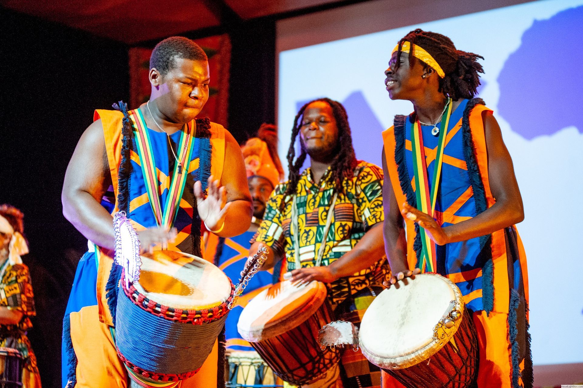 African drummers in vibrant blue and orange outfits, playing drums on a stage.