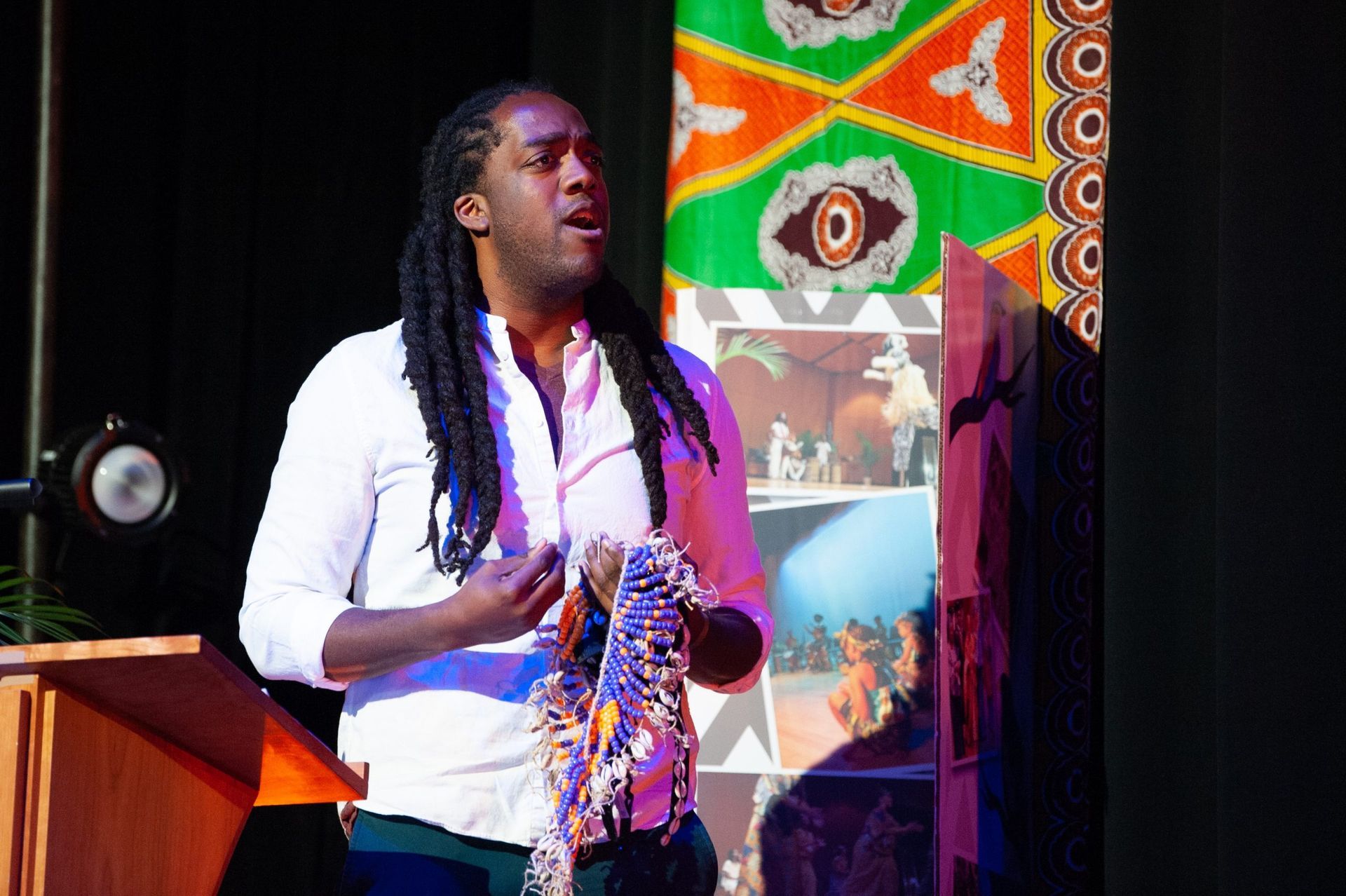 Man with dreadlocks speaking at a podium, holding fabric. Background includes colorful textiles and artwork.