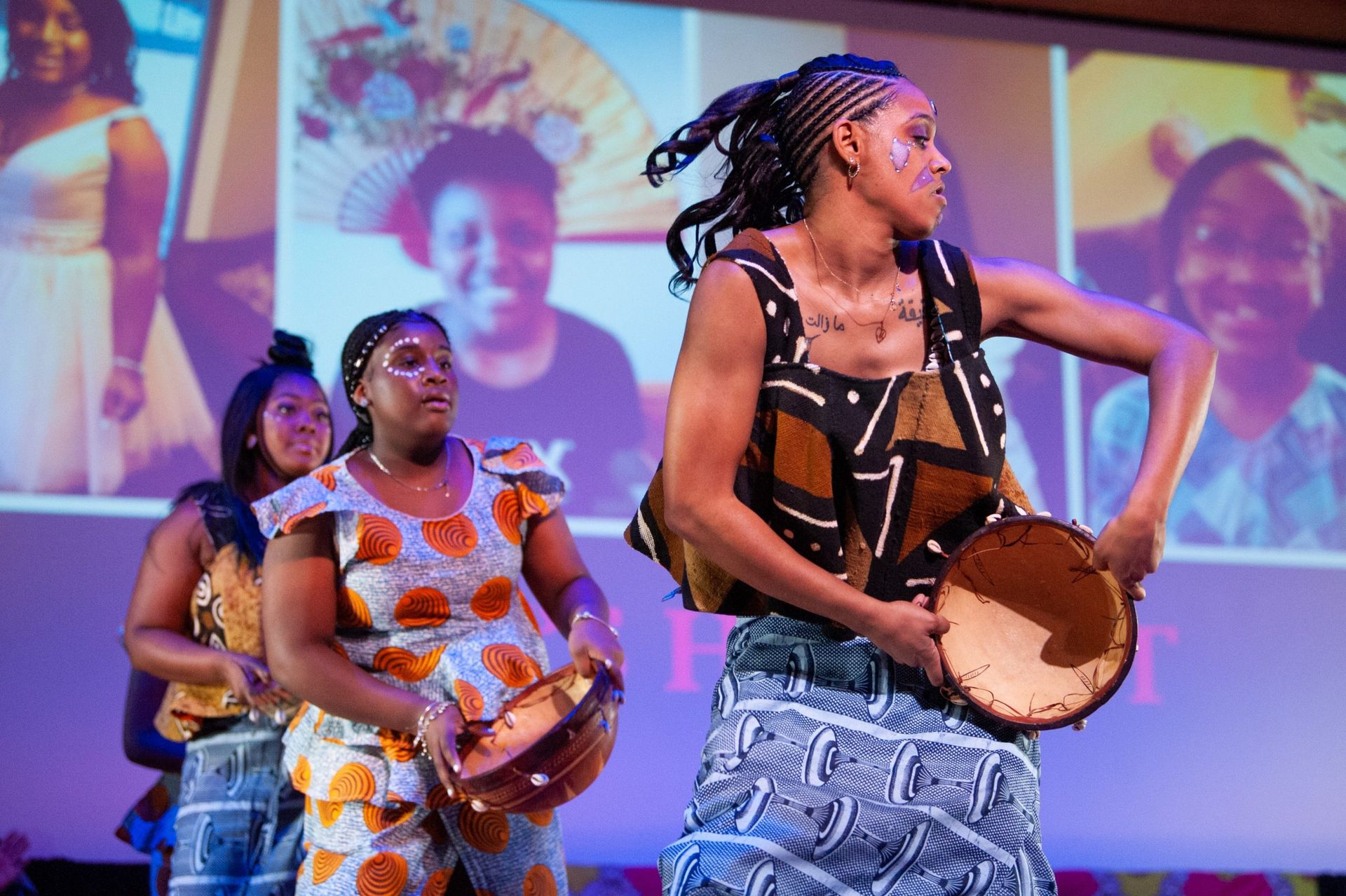 Women in colorful African clothing dance and play drums on stage. Background shows smiling women.
