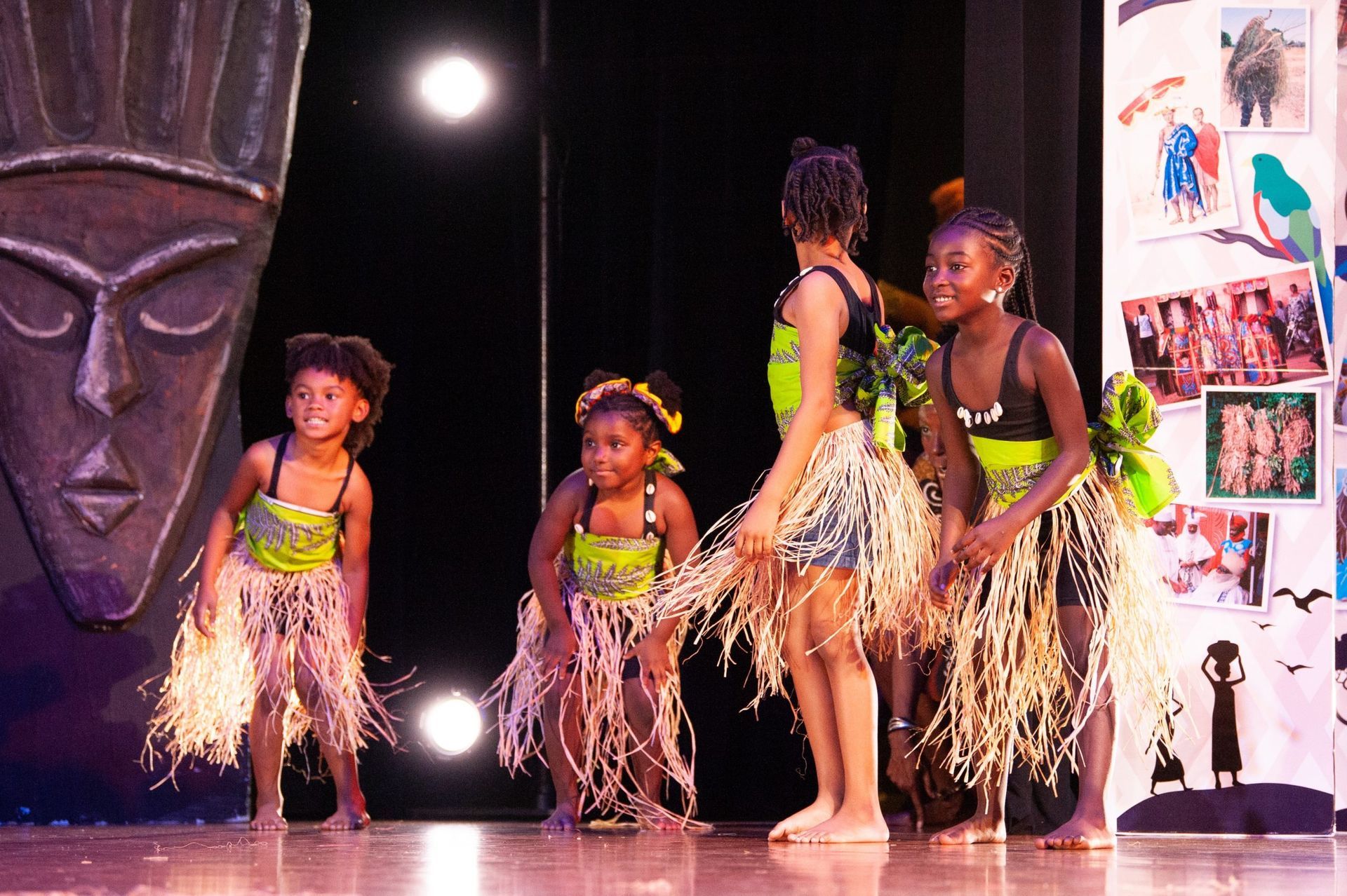 Young girls in grass skirts dance on a stage with an African mask backdrop.