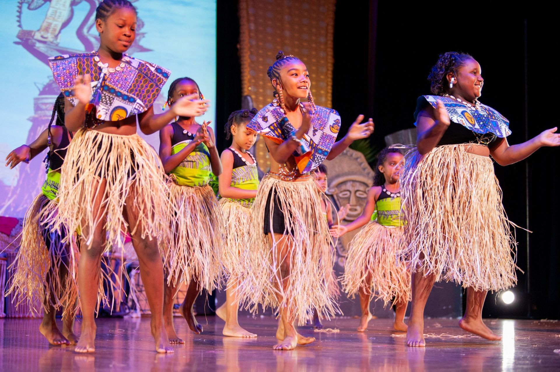 Group of children dancing on stage in grass skirts and colorful tops.