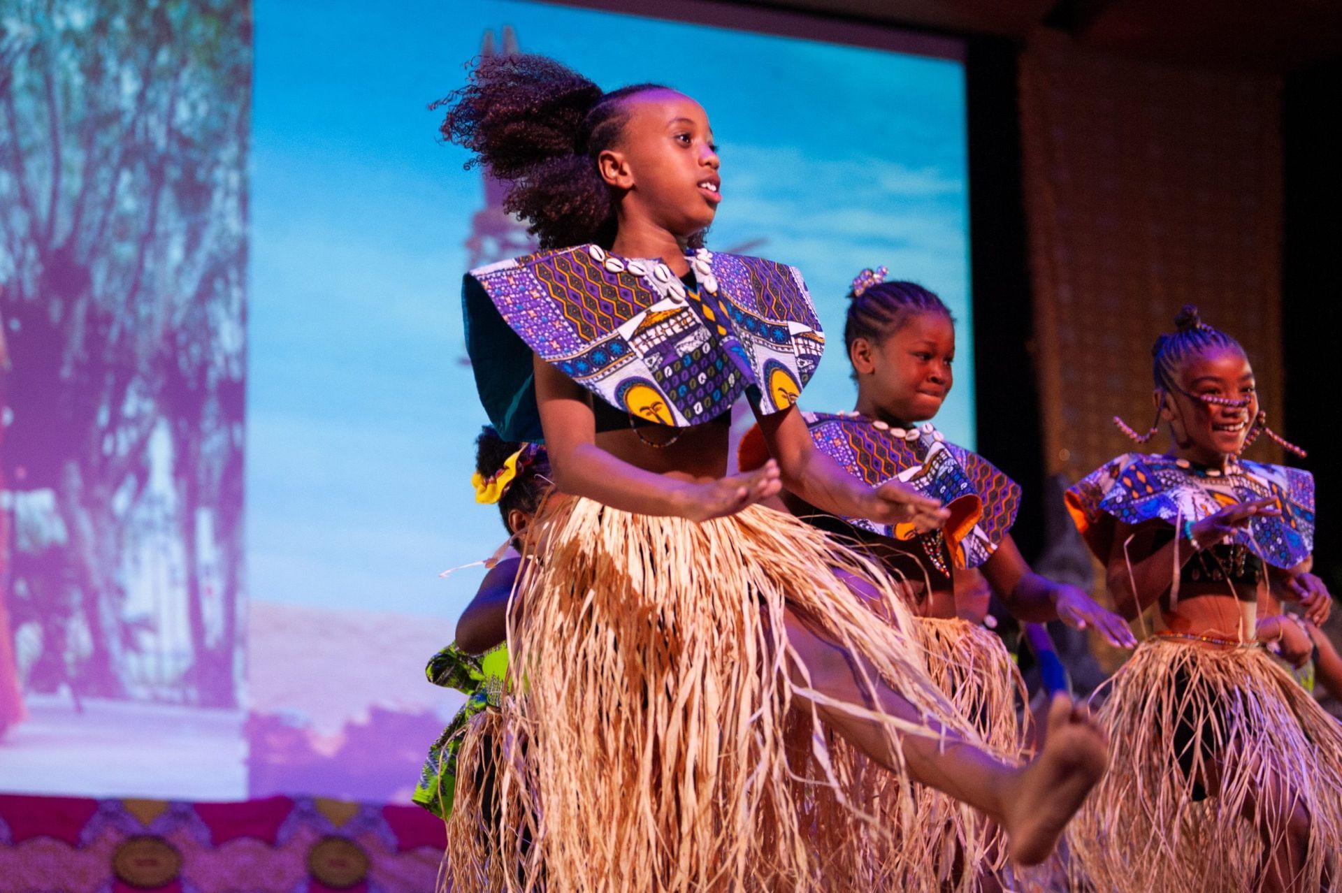 Three young girls in African attire dance on stage, with smiles and energetic movements.