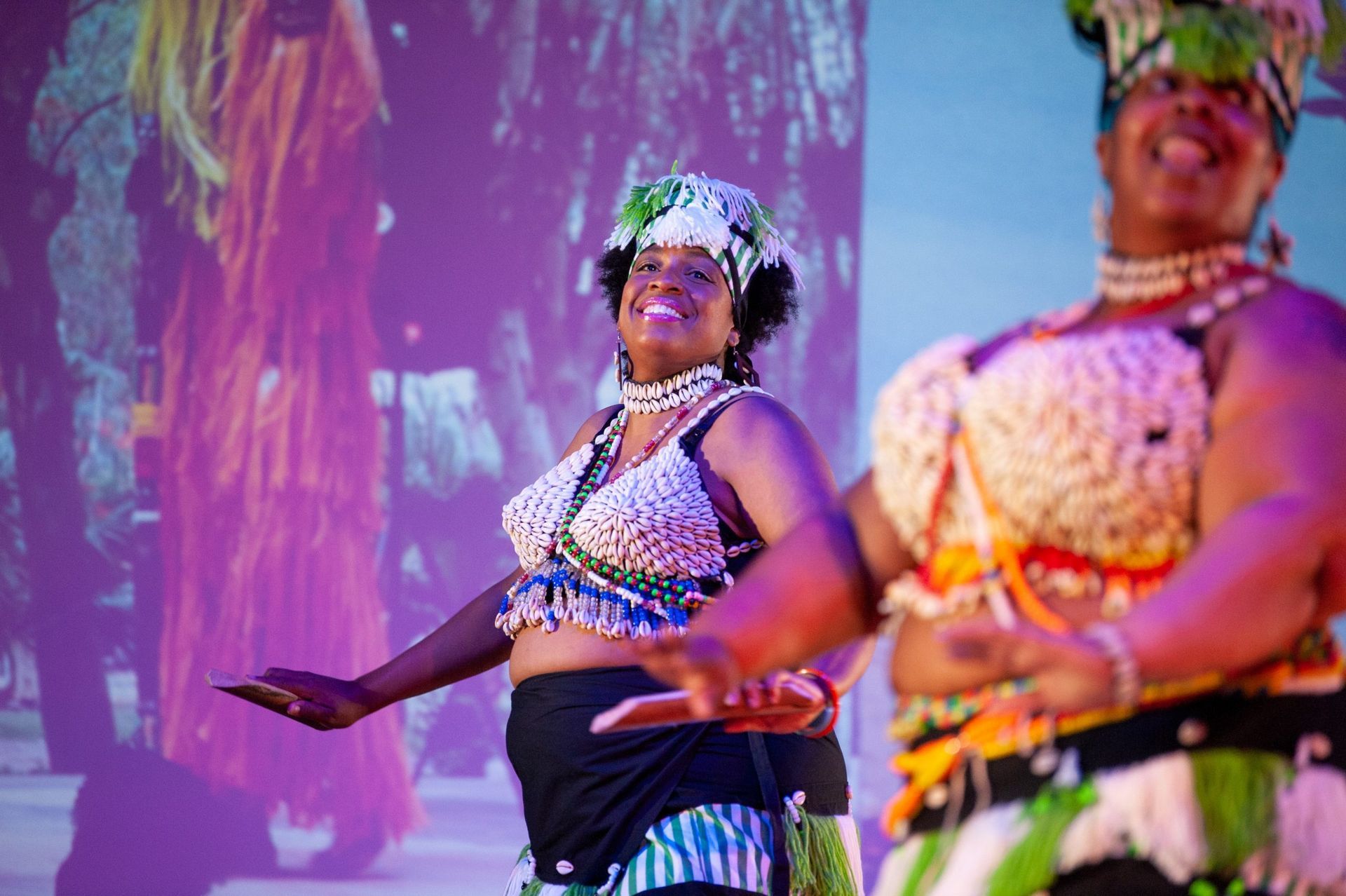 Two women in colorful beaded attire dance, smiling in front of a projected background.