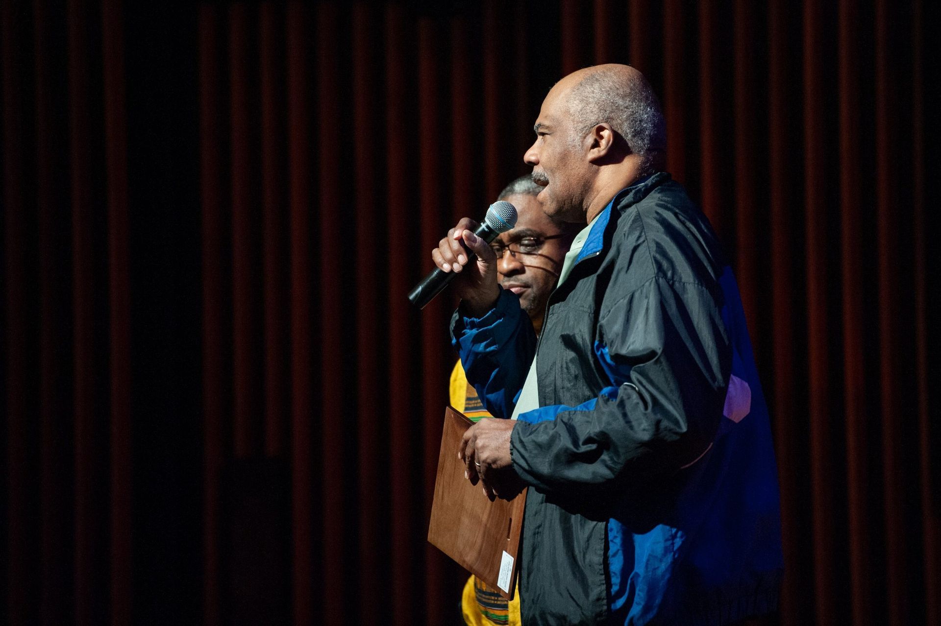 Two men onstage with microphones, one holding a clipboard. Dark stage with red curtains.