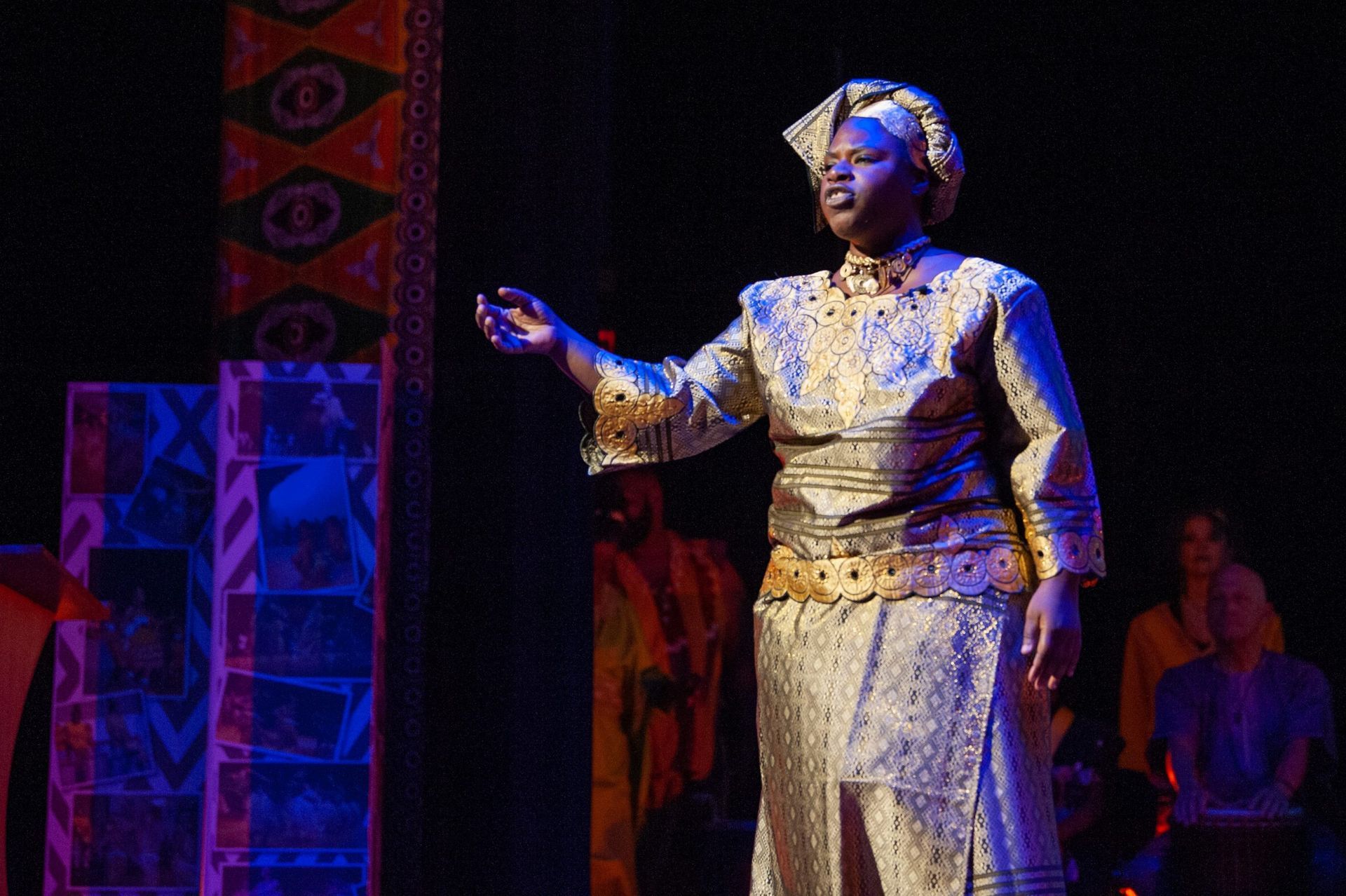 Woman in gold costume on stage, gesturing. Dark setting with audience in background.