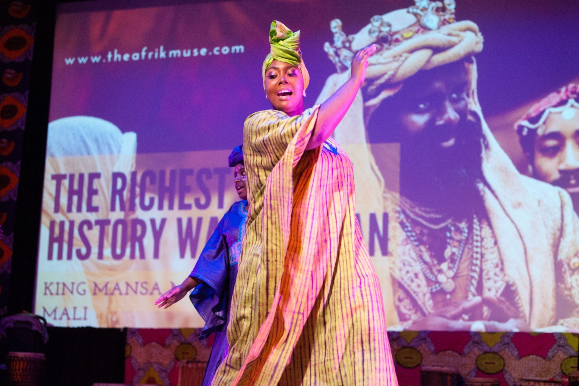 Woman in African attire on stage, arms raised, with projection of King Mansa Musa, purple lighting.
