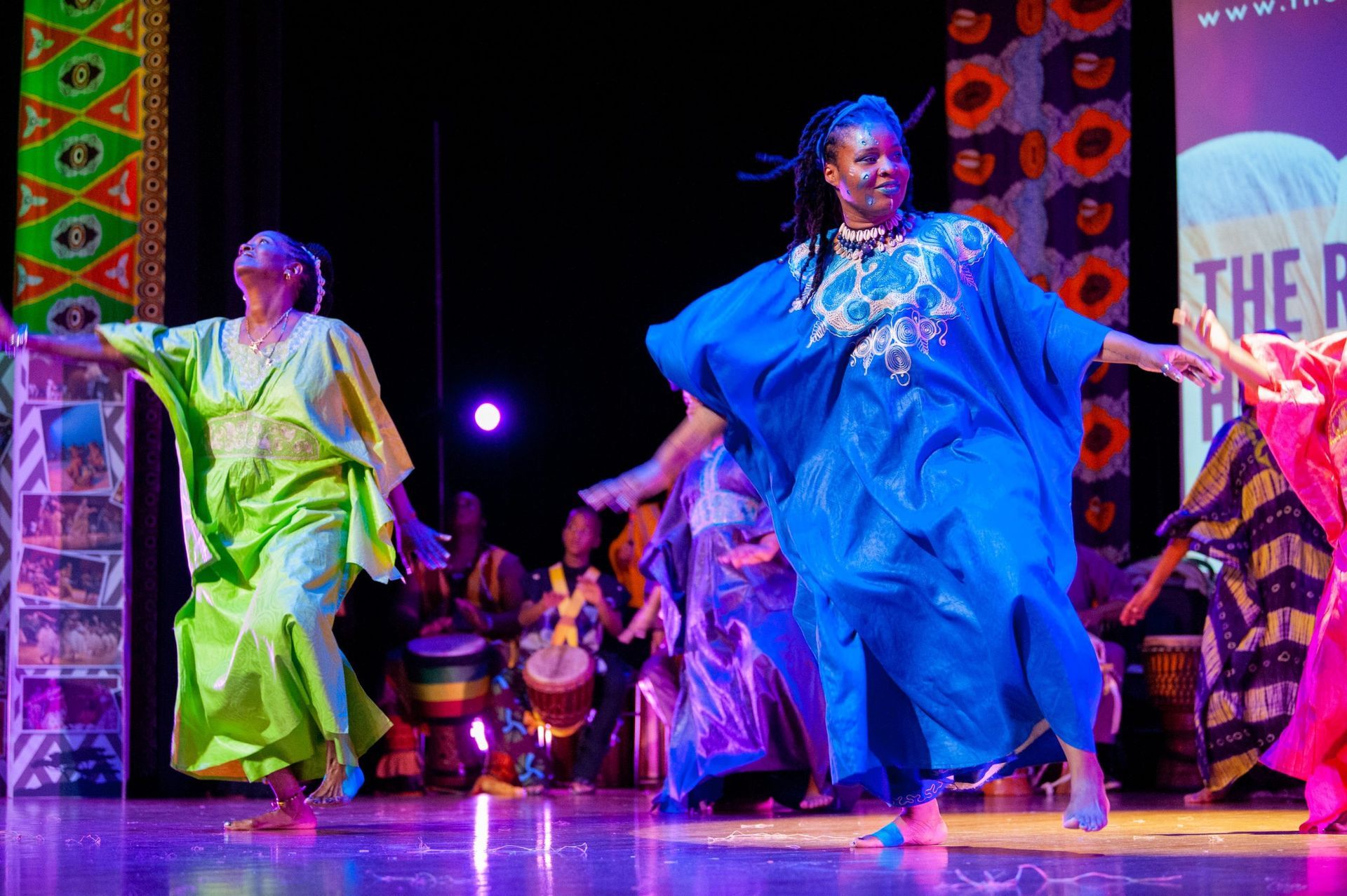 Dancers in colorful robes on stage, performing. Women in green, blue, and pink, barefoot. Drums in background.