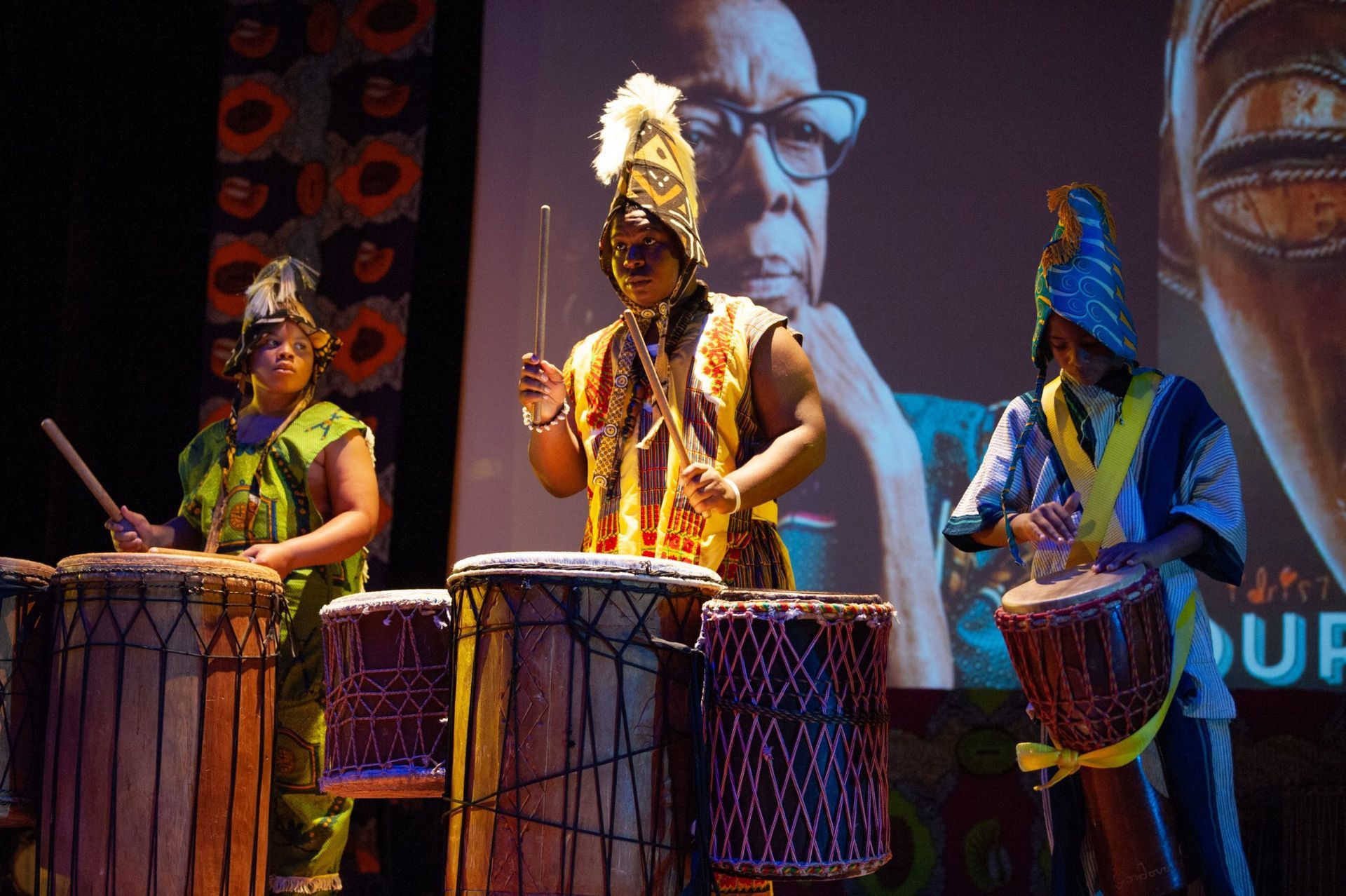 Three drummers in African attire perform onstage, with a portrait projected behind them.
