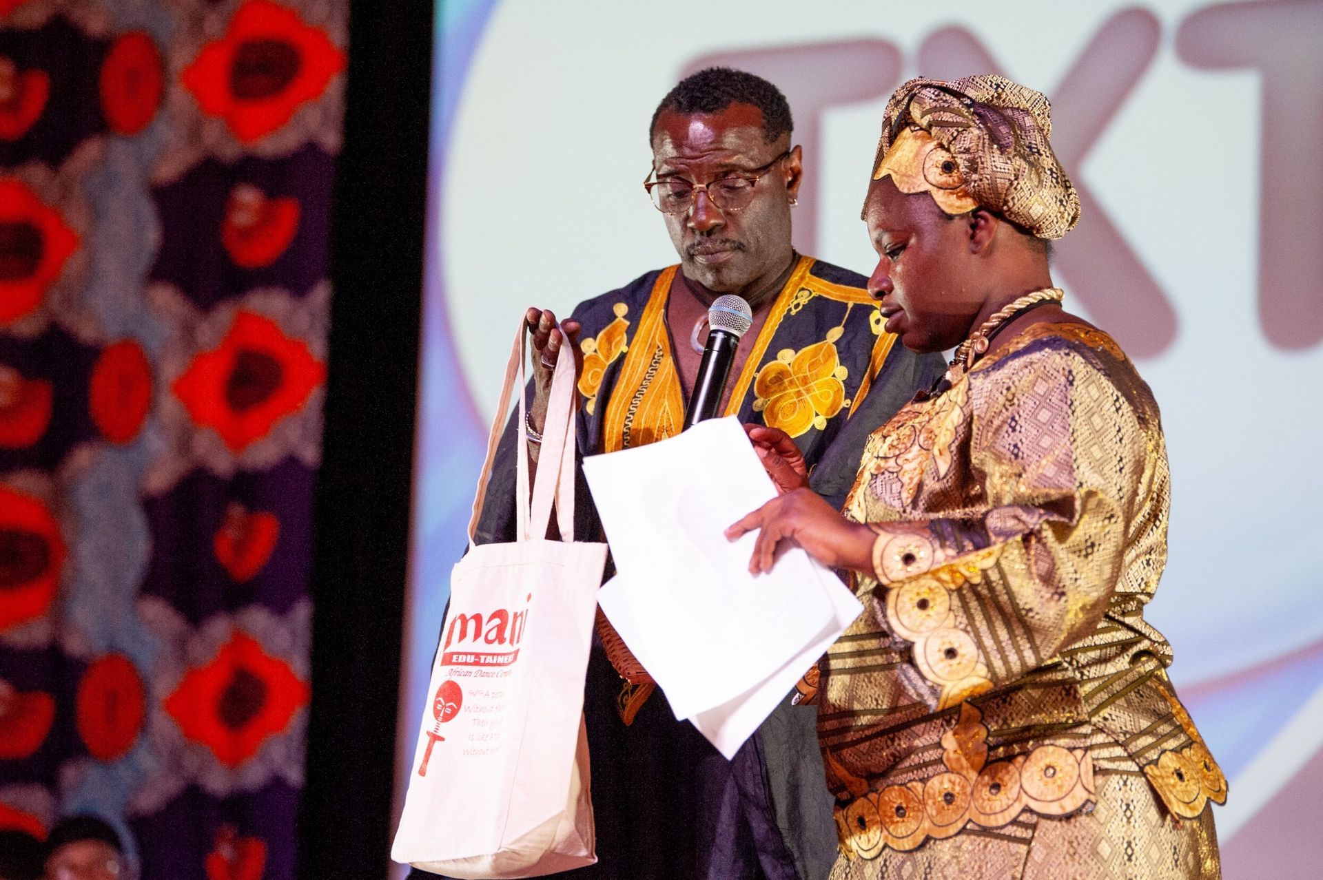 Two people on stage, one holding a tote bag, reviewing papers. Floral backdrop, warm lighting.