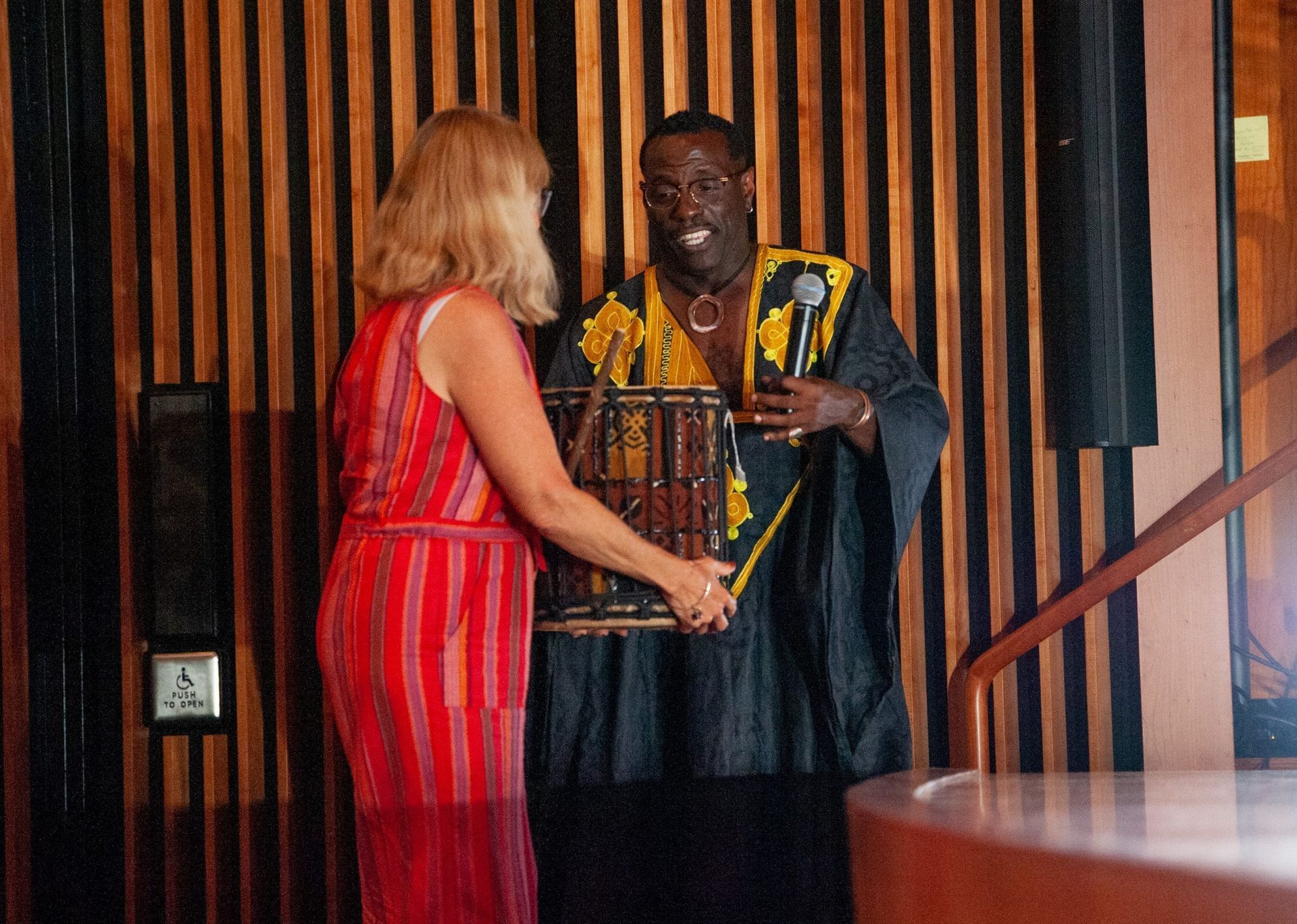 Woman and man with a drum in front of a wood wall, the man in traditional clothes, likely at an event.