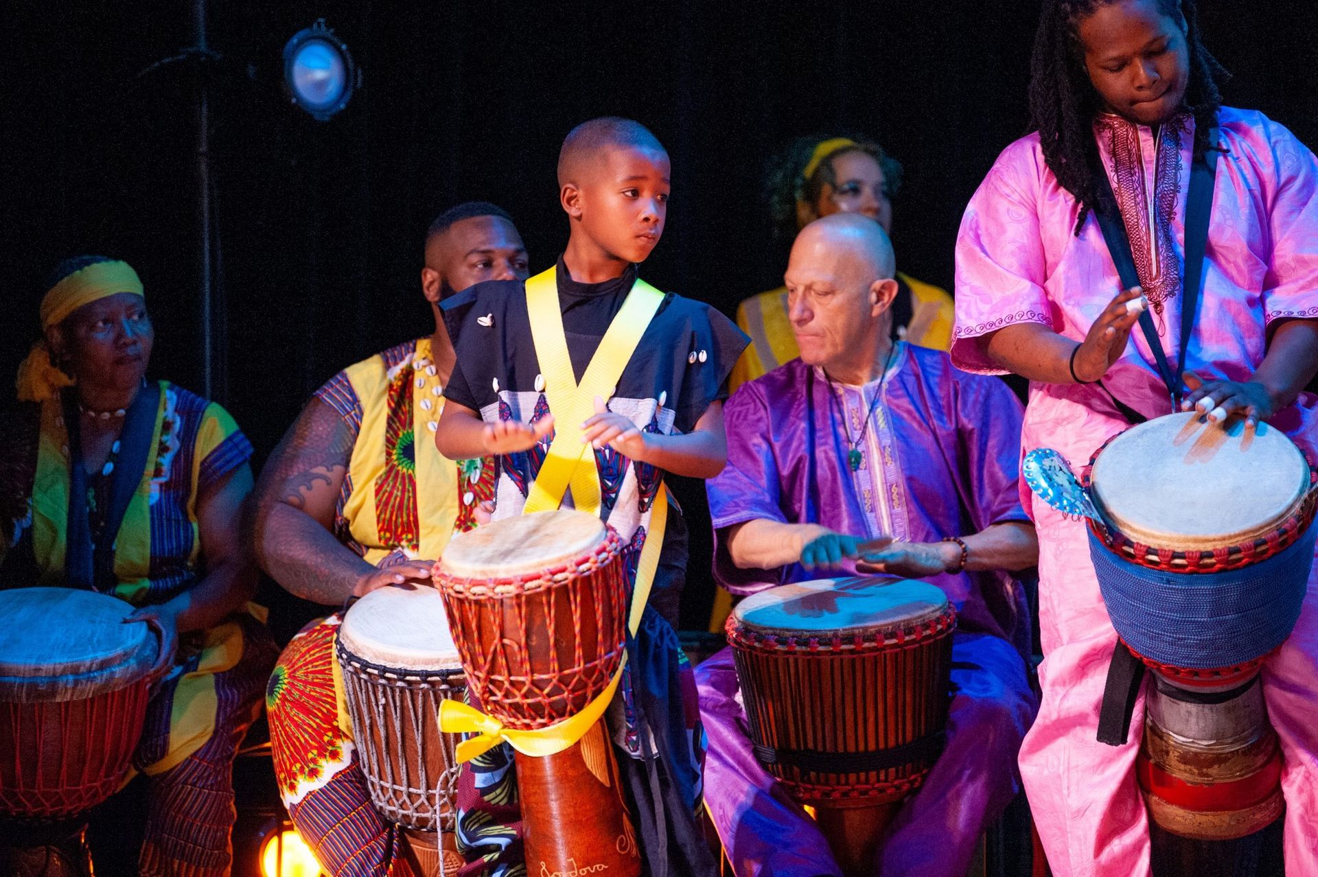 Group of people playing drums, stage lit, wearing bright colors, focused expressions.