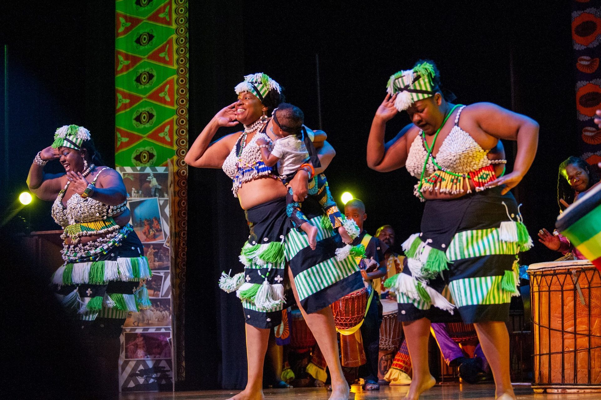 Women in colorful costumes dancing onstage, one holding a baby, with drummers.