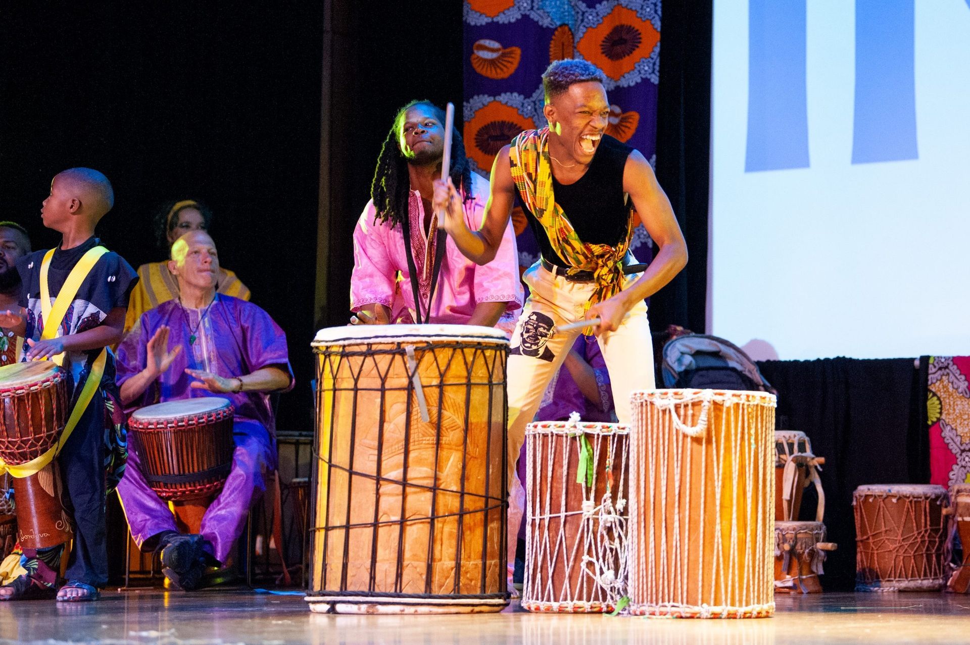 A group of people playing African drums on a stage; one person enthusiastically drumming.
