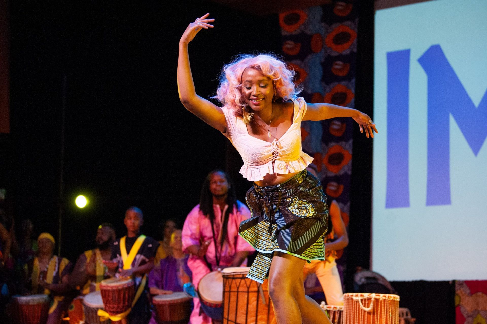 A woman with pink hair dances onstage, a drum circle behind her.