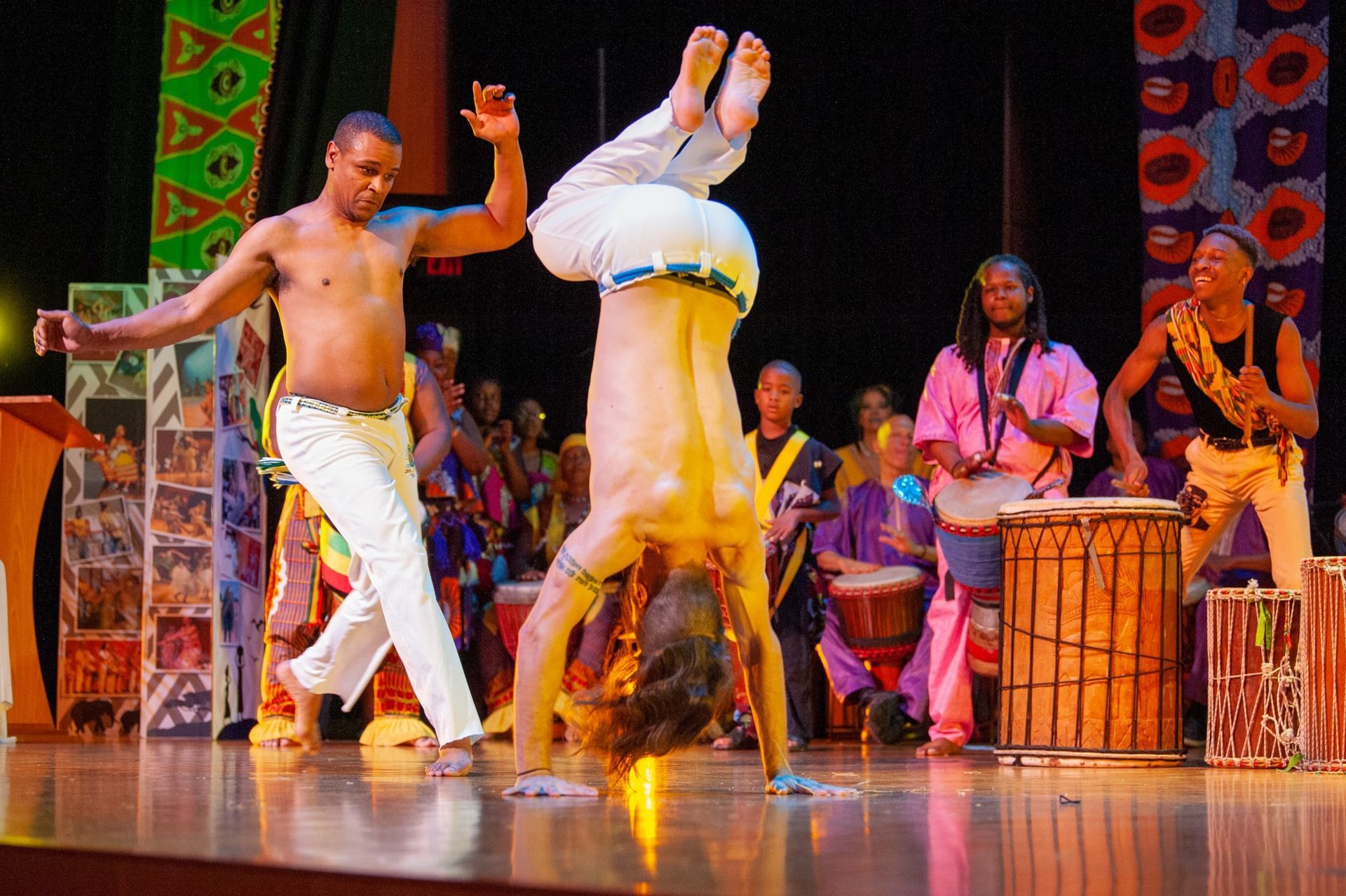 Men performing Capoeira moves on stage, one inverted in a handstand. Drummers, colorful backdrop.
