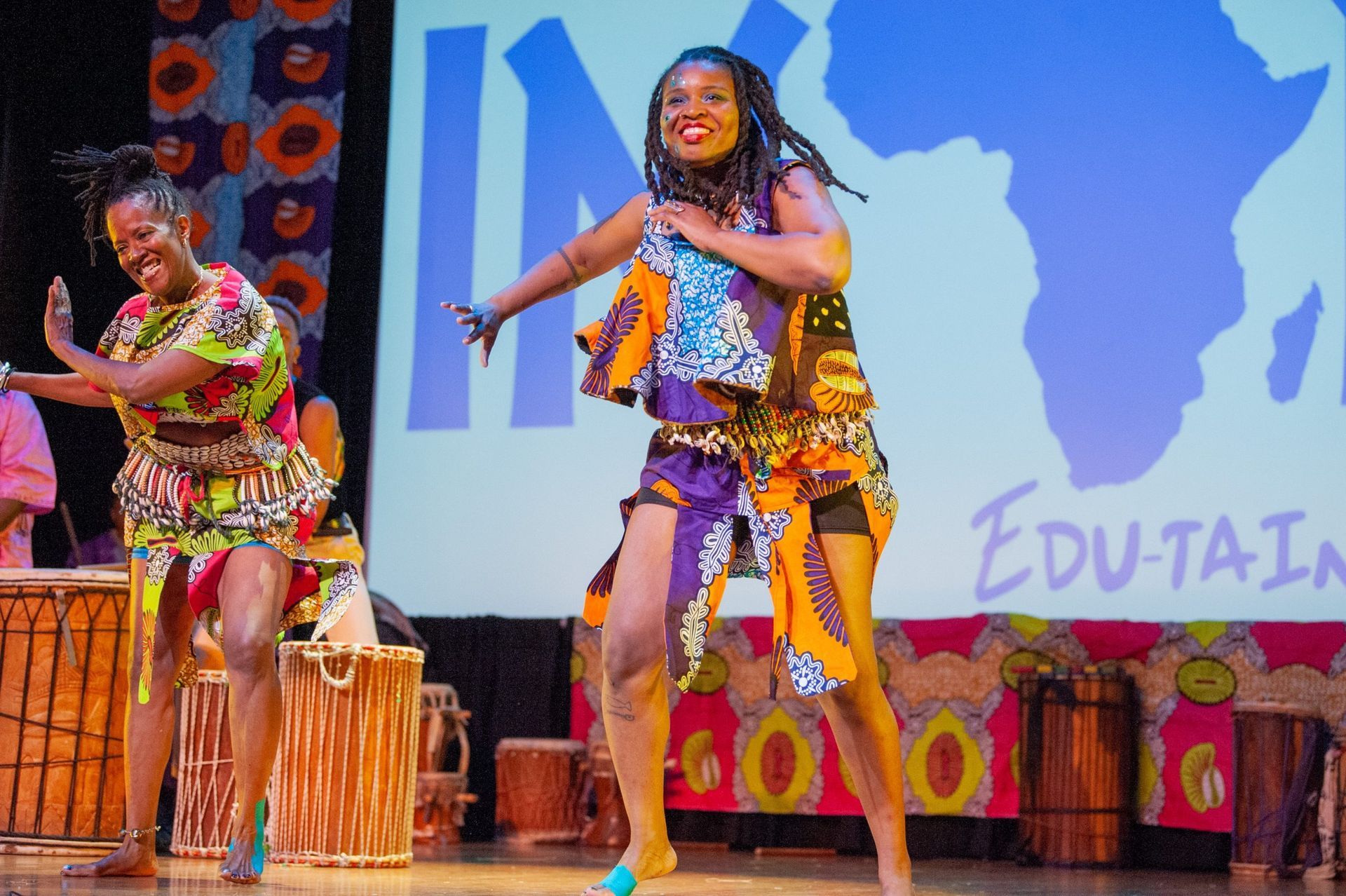 Two women dancing onstage in colorful African attire, with drums and an African map backdrop.