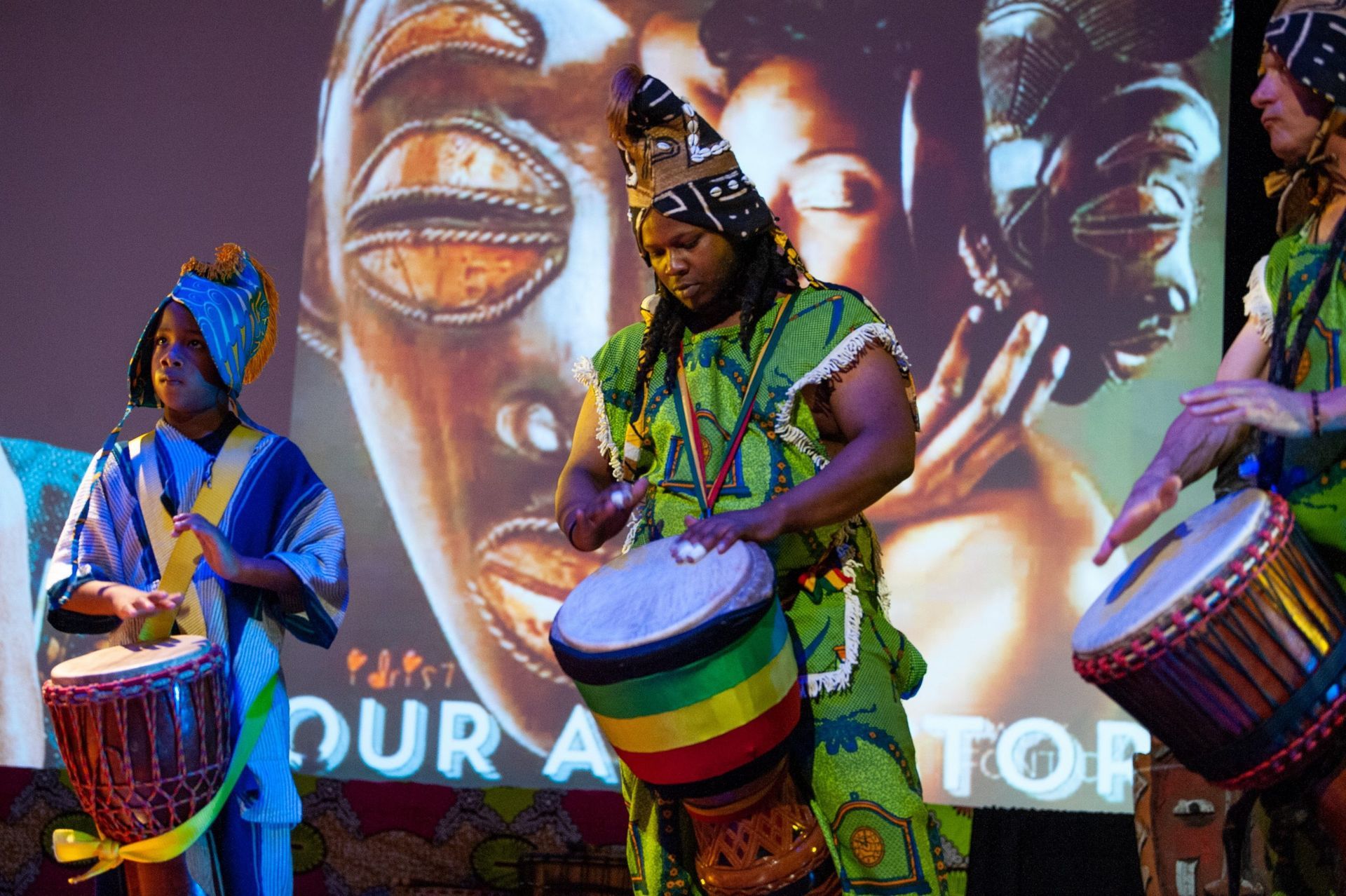 People in African attire playing drums on stage, backdrop of African art.