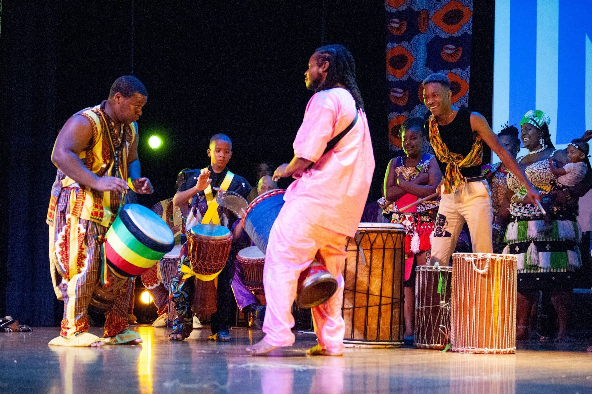 African drummers on stage; men and youths playing drums, dancing, vibrant colors.