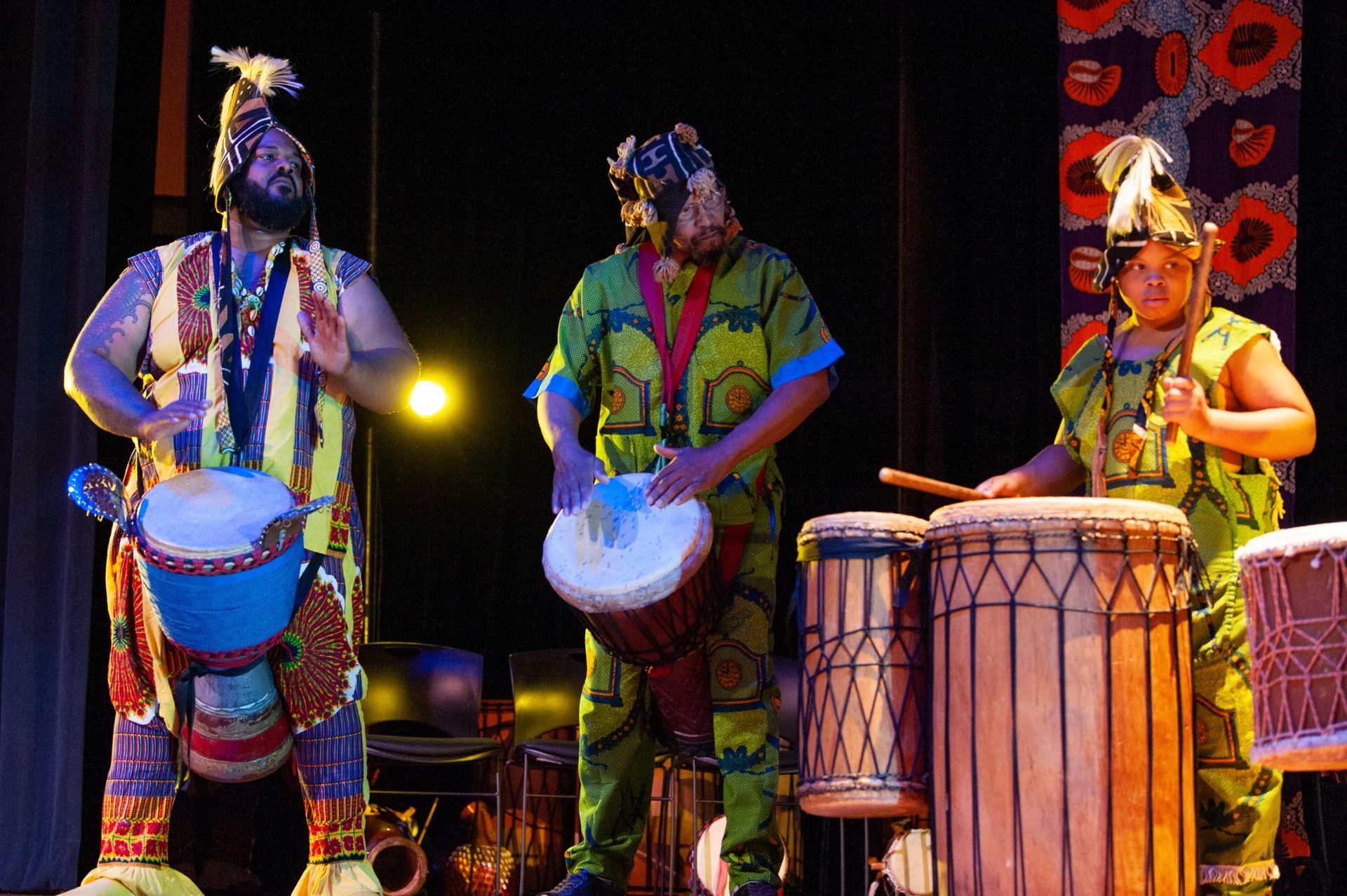 Three people in colorful African attire playing drums on a stage with a dark background.