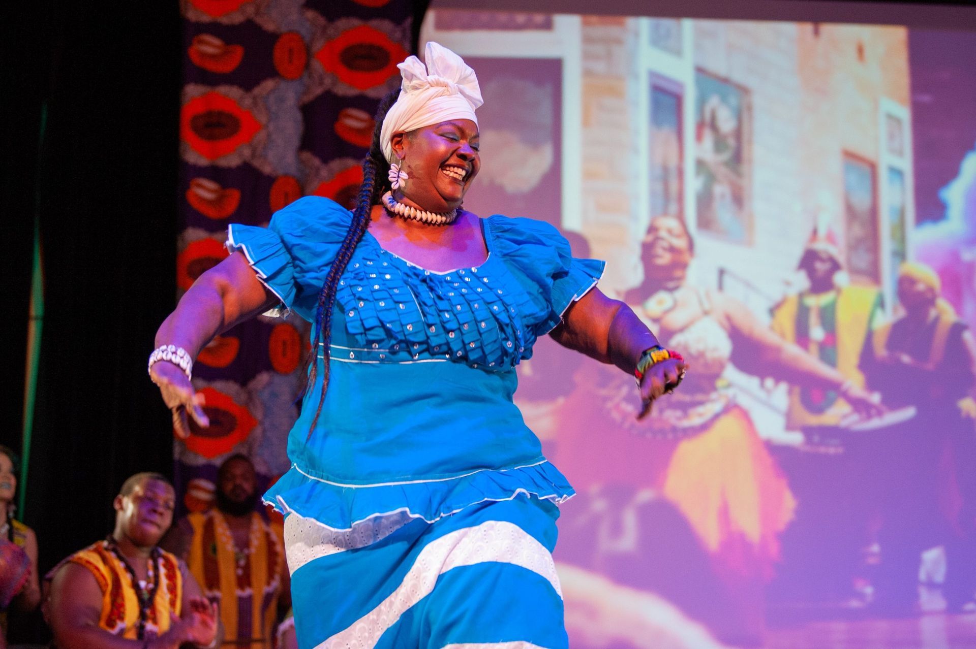 Woman in blue dress dancing on stage, smiling, background projection of other dancers.