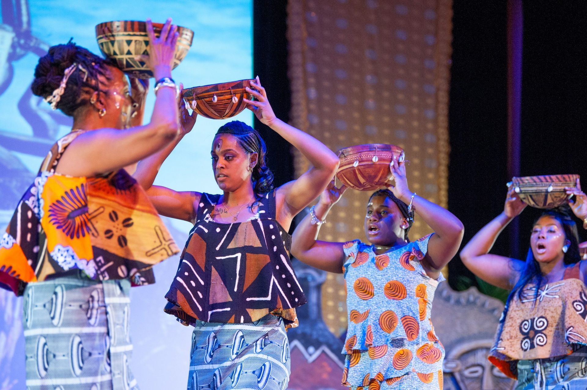Women in colorful African attire balancing bowls on their heads, performing on stage.