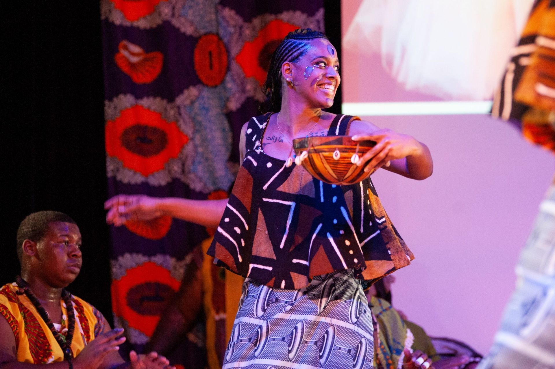 Woman in African print dress dances on stage, holding a wooden bowl.