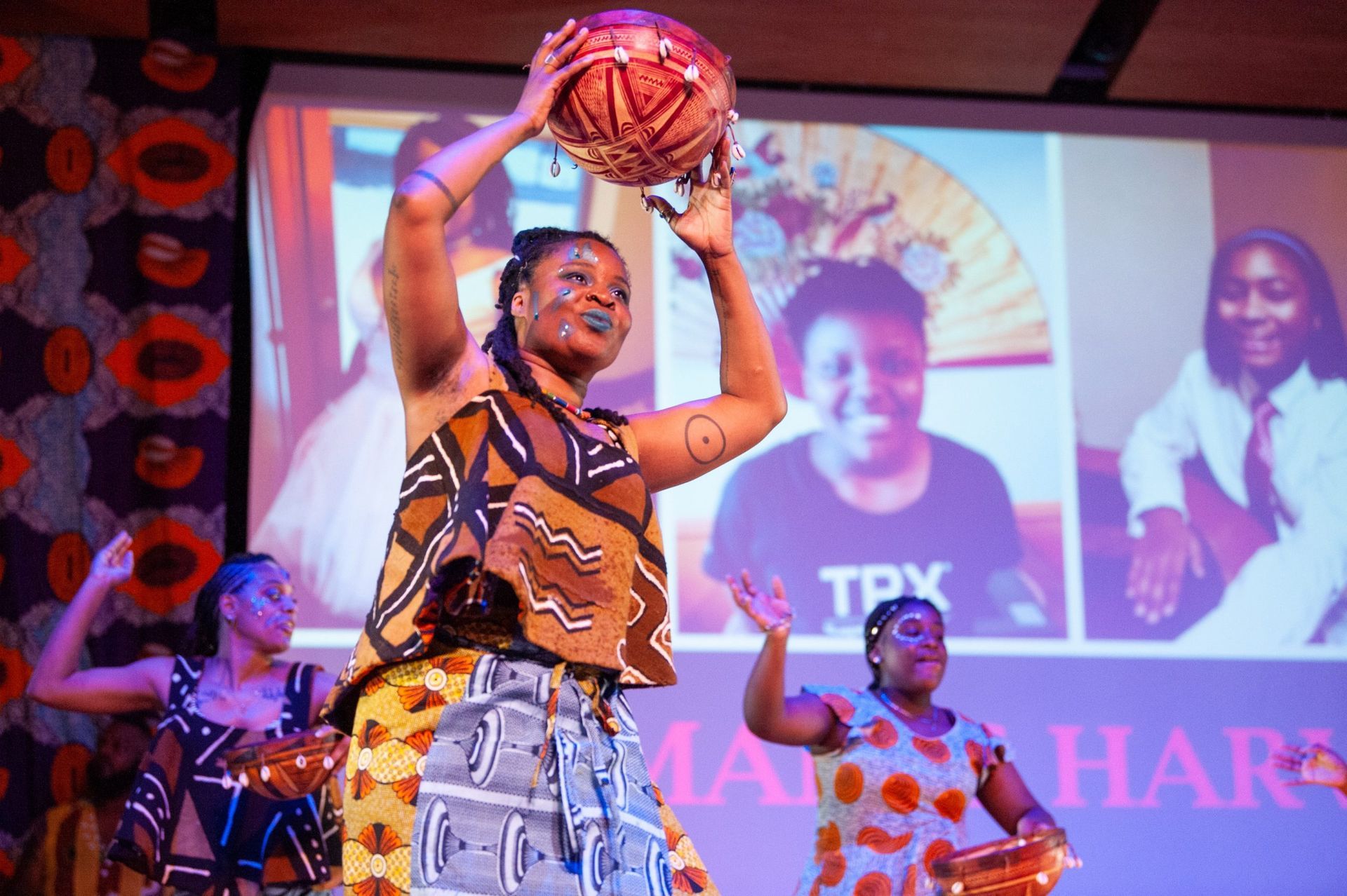 Dancers in African attire perform onstage, one holding a woven ball aloft, with projected images behind them.