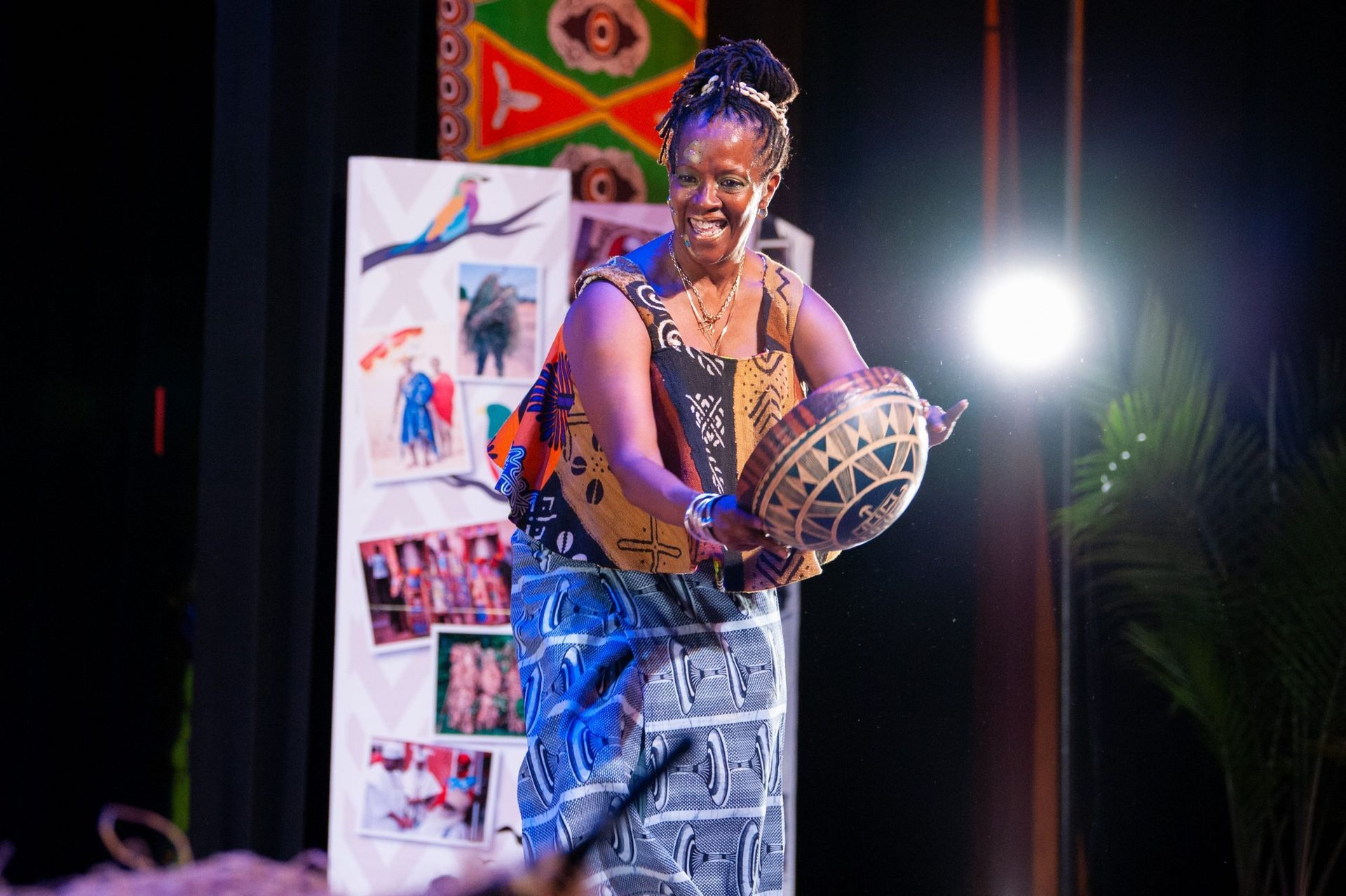 Woman in African attire holds a decorated gourd on stage. Smiling, in front of a decorated backdrop, spotlight.