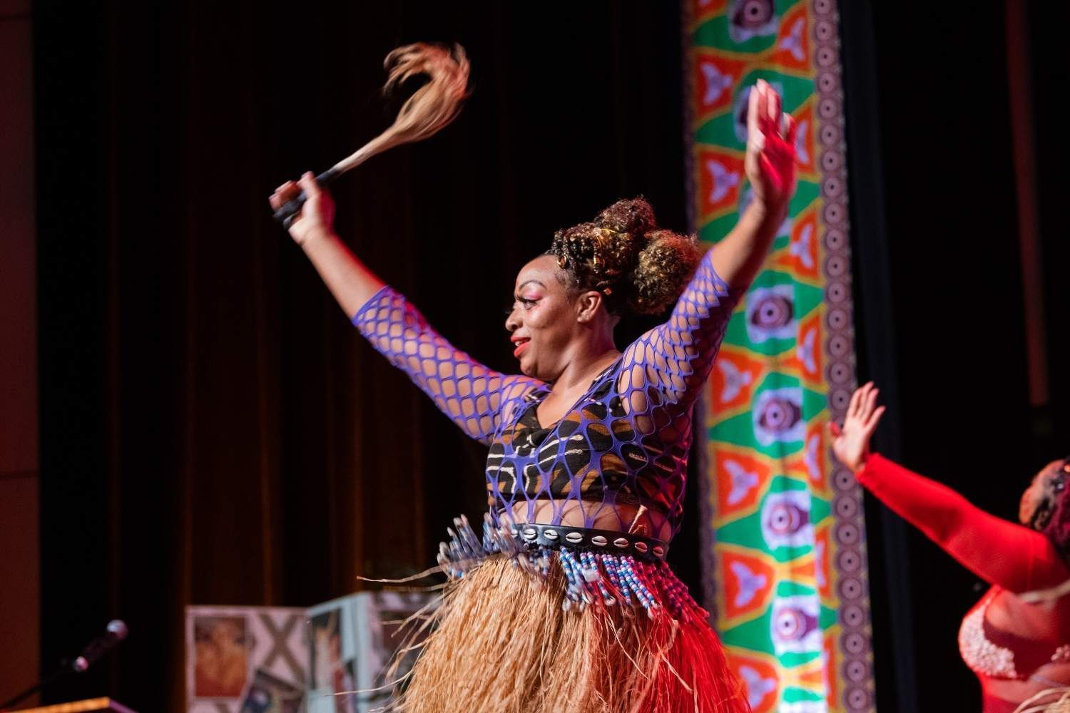 Woman in colorful costume dancing on stage, holding a feather fan.