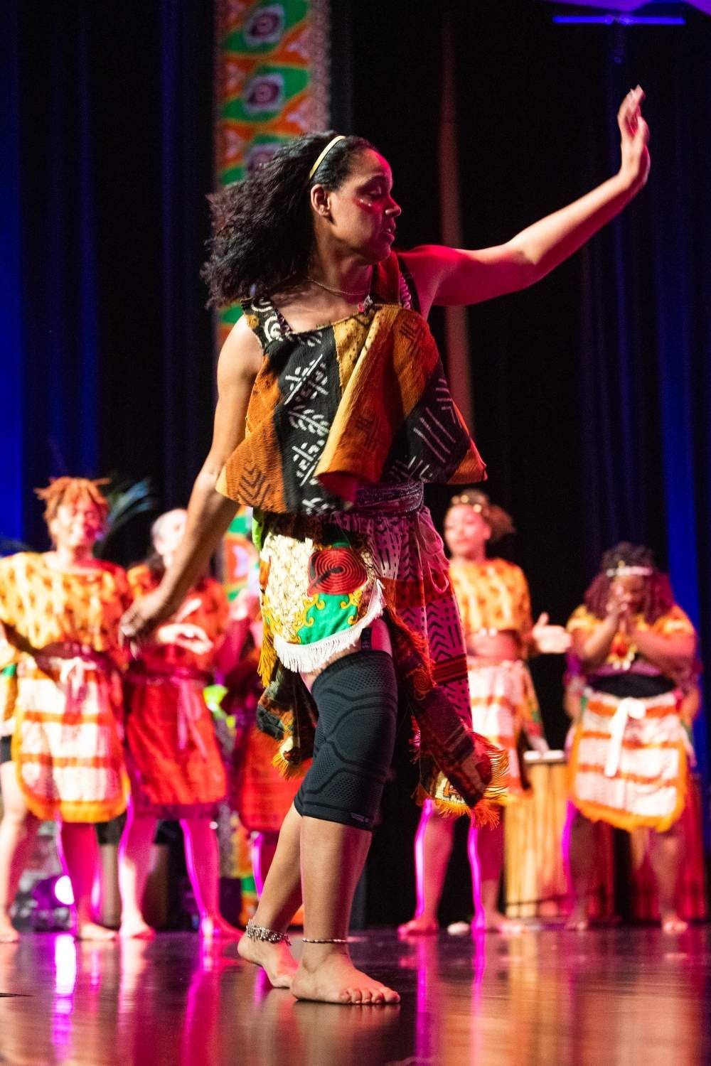 Woman in patterned costume dancing on stage; other dancers in background.