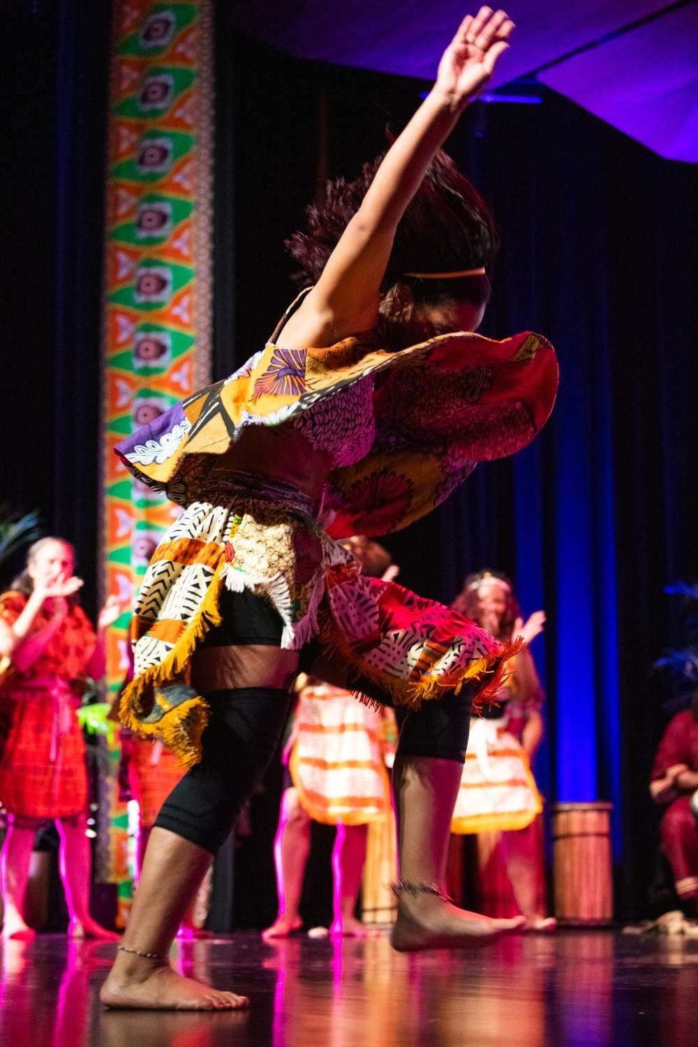 A person dancing in a colorful costume on stage with a group of dancers in the background.