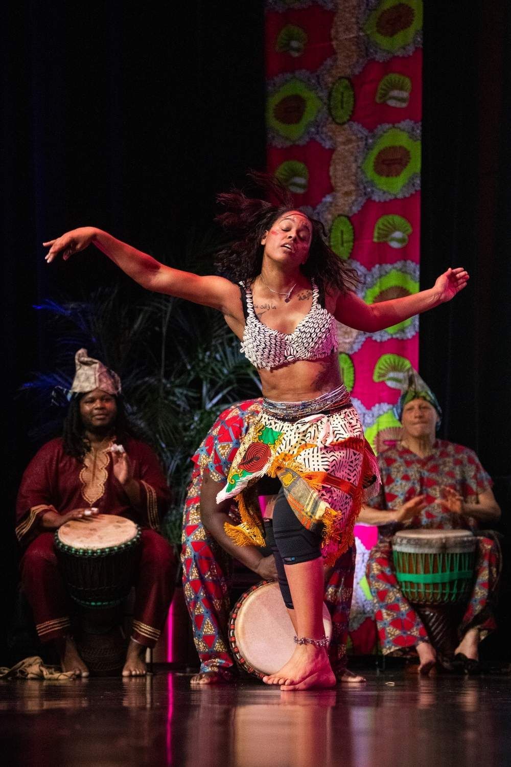 African dancer in a beaded top and skirt, performing with drummers on stage.