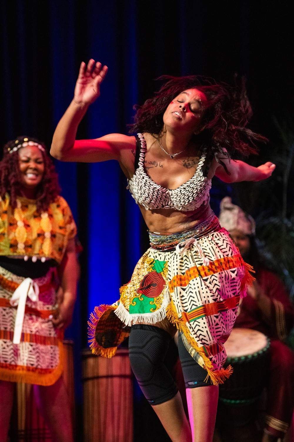 Woman dancing on stage in colorful African attire. Another woman smiles in background.