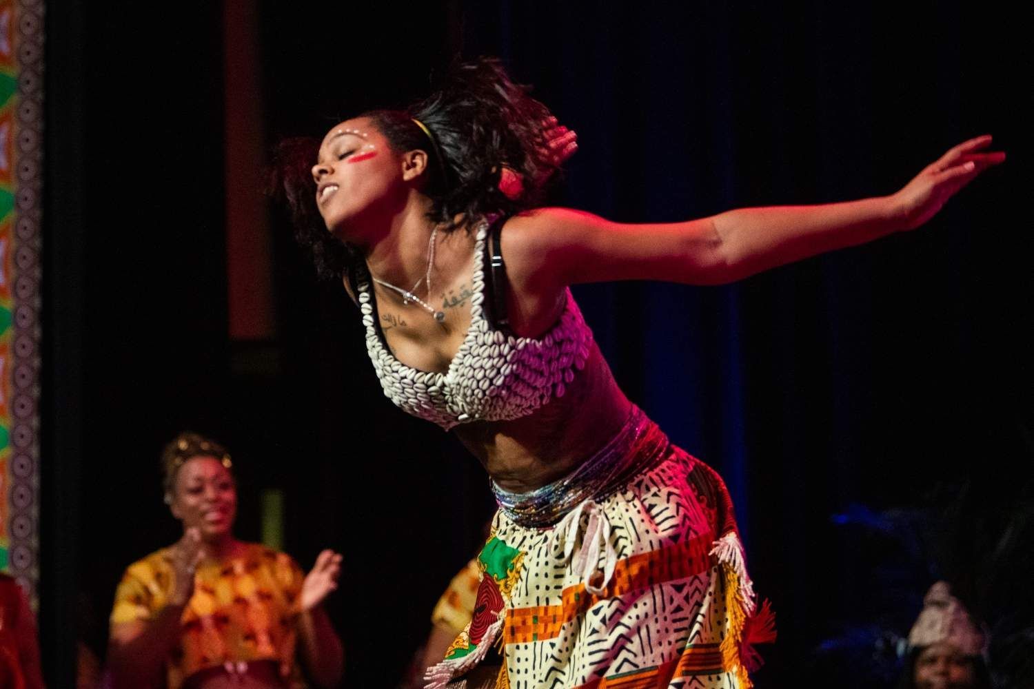 Woman dancing onstage, wearing a white beaded top and patterned skirt, arms outstretched, joyful expression.
