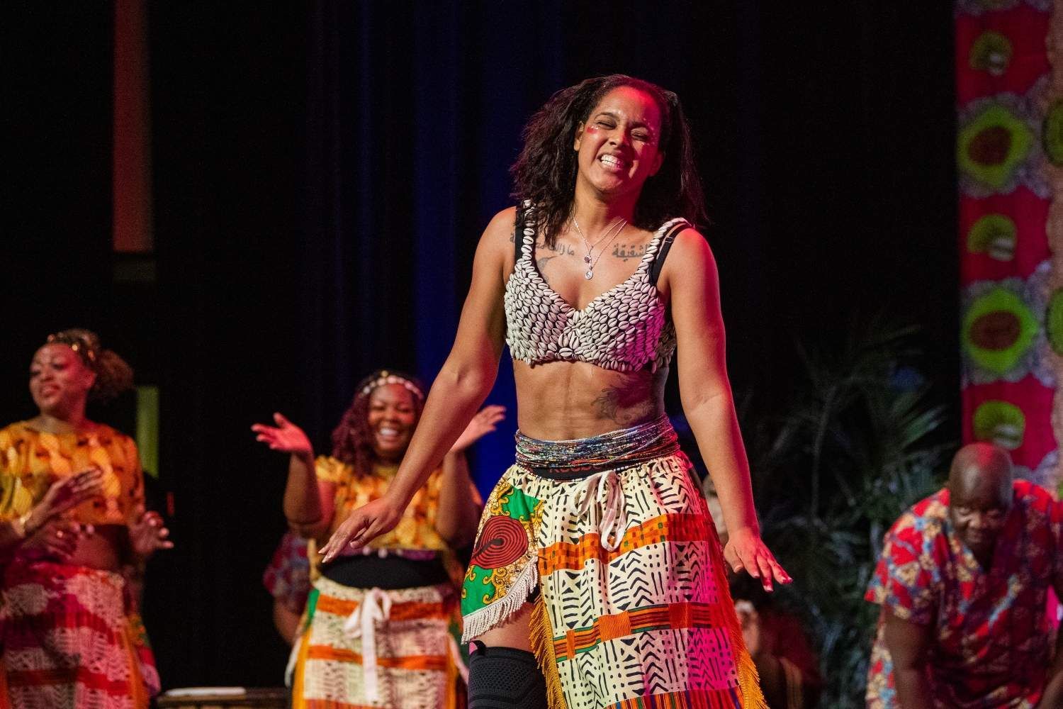 Woman dancing onstage, wearing a decorated crop top and patterned skirt, smiling. Other performers in colorful outfits.