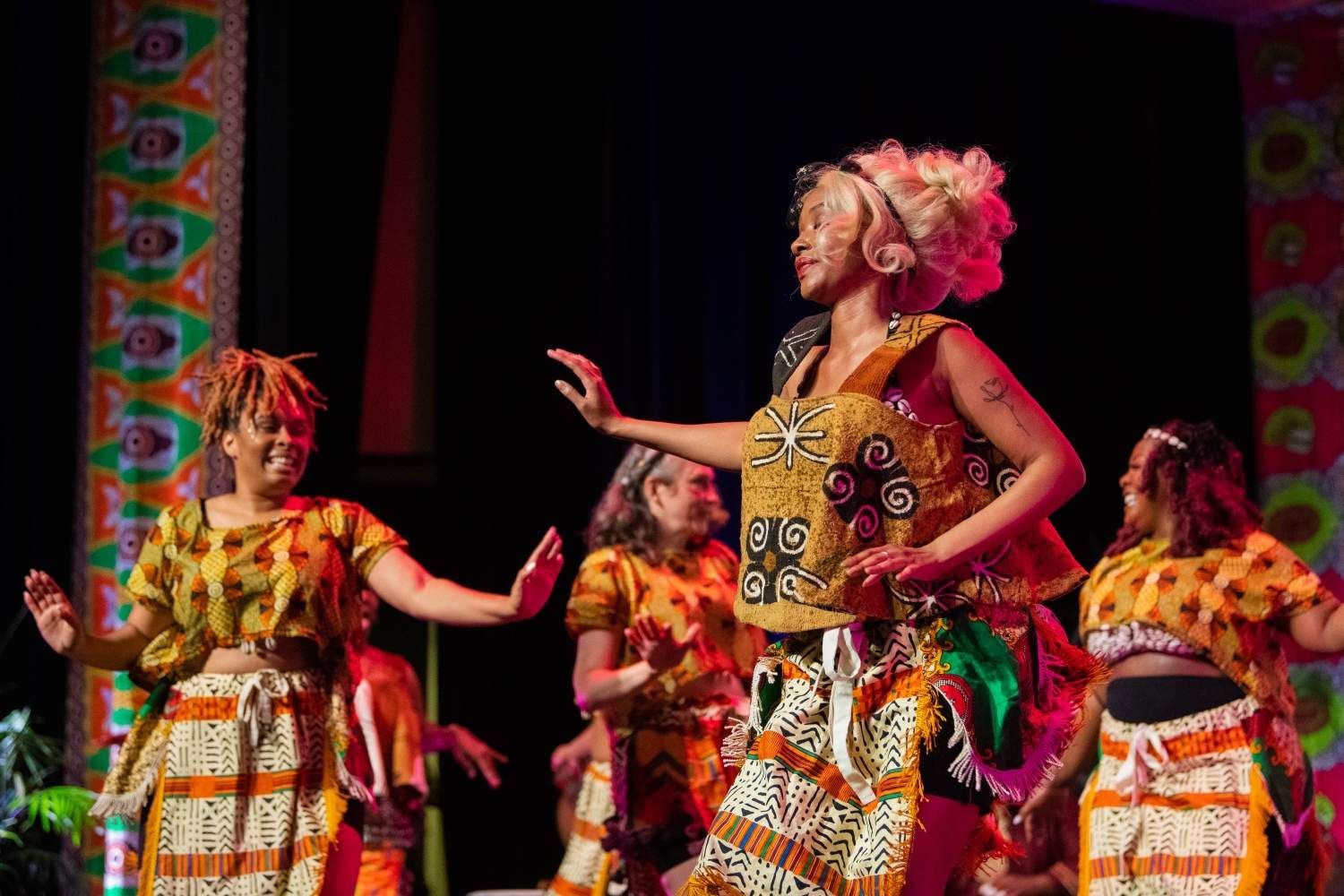 Group of women dancing on stage, wearing colorful patterned outfits.