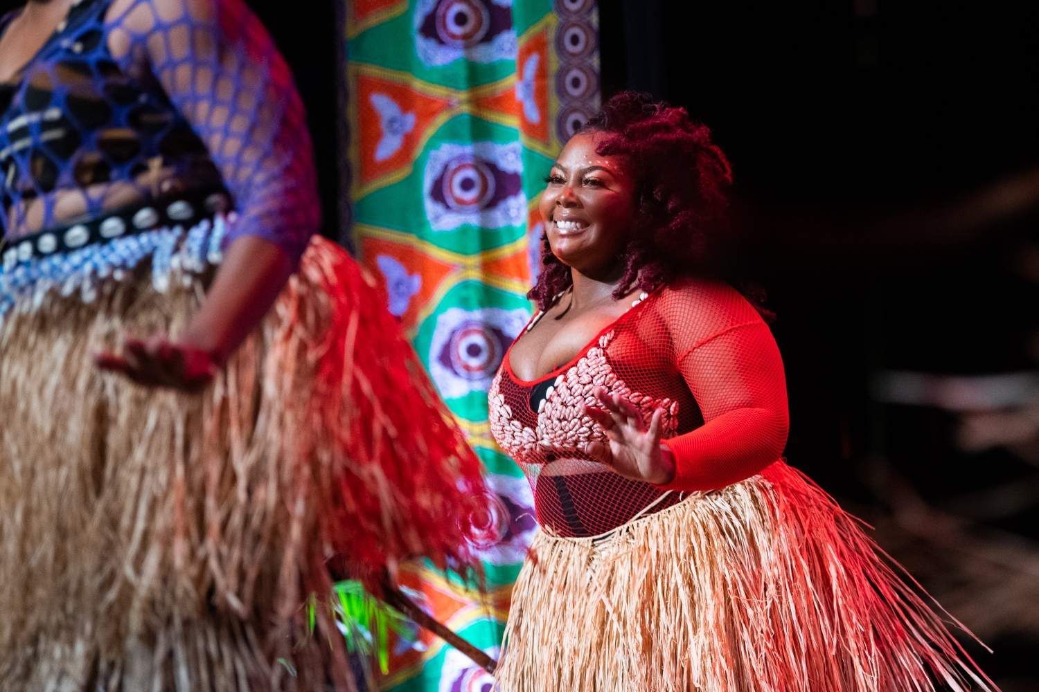 Woman in red ensemble dances on stage with a straw skirt, smiling, with colorful backdrop.