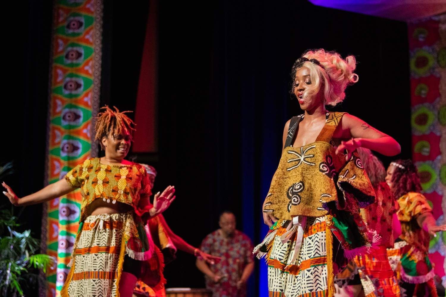 Two women in colorful African-style clothing dance on a stage with decorated pillars.
