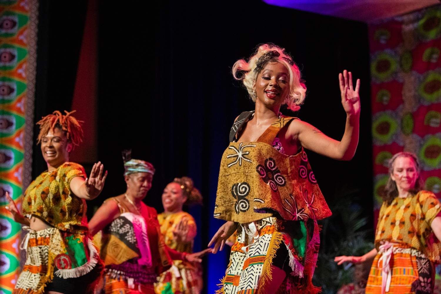 Dancers in colorful African print costumes perform onstage, arms raised, smiling.