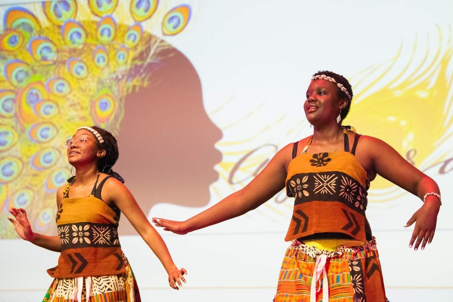 Two women dance on stage in front of a backdrop with peacock feathers and a silhouette.