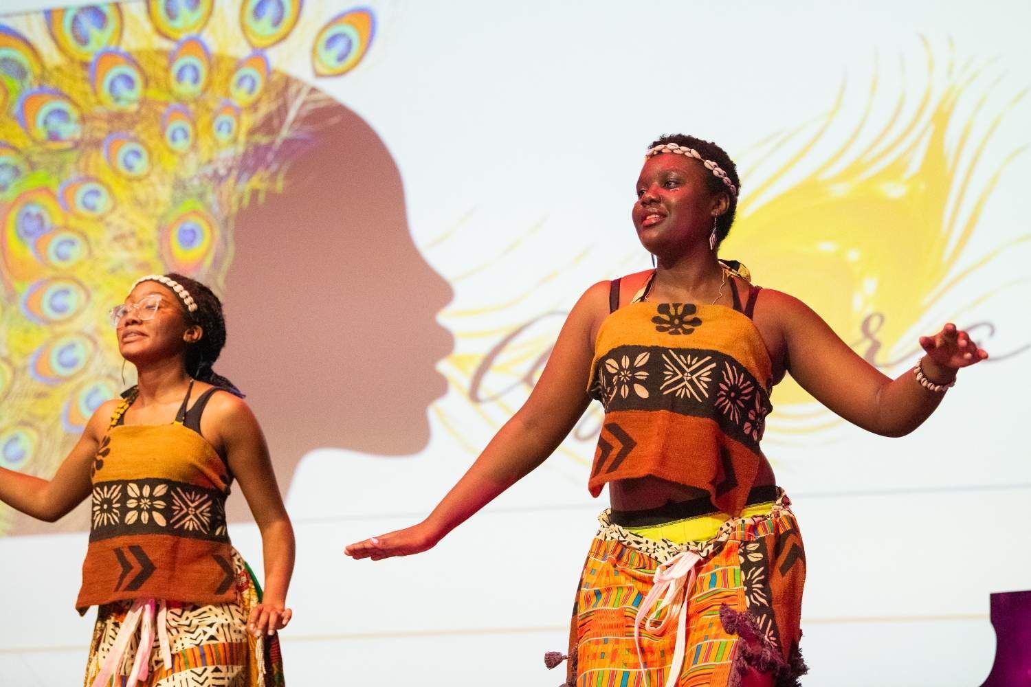 Two women in African attire dance on stage with a peacock feather and silhouette backdrop.