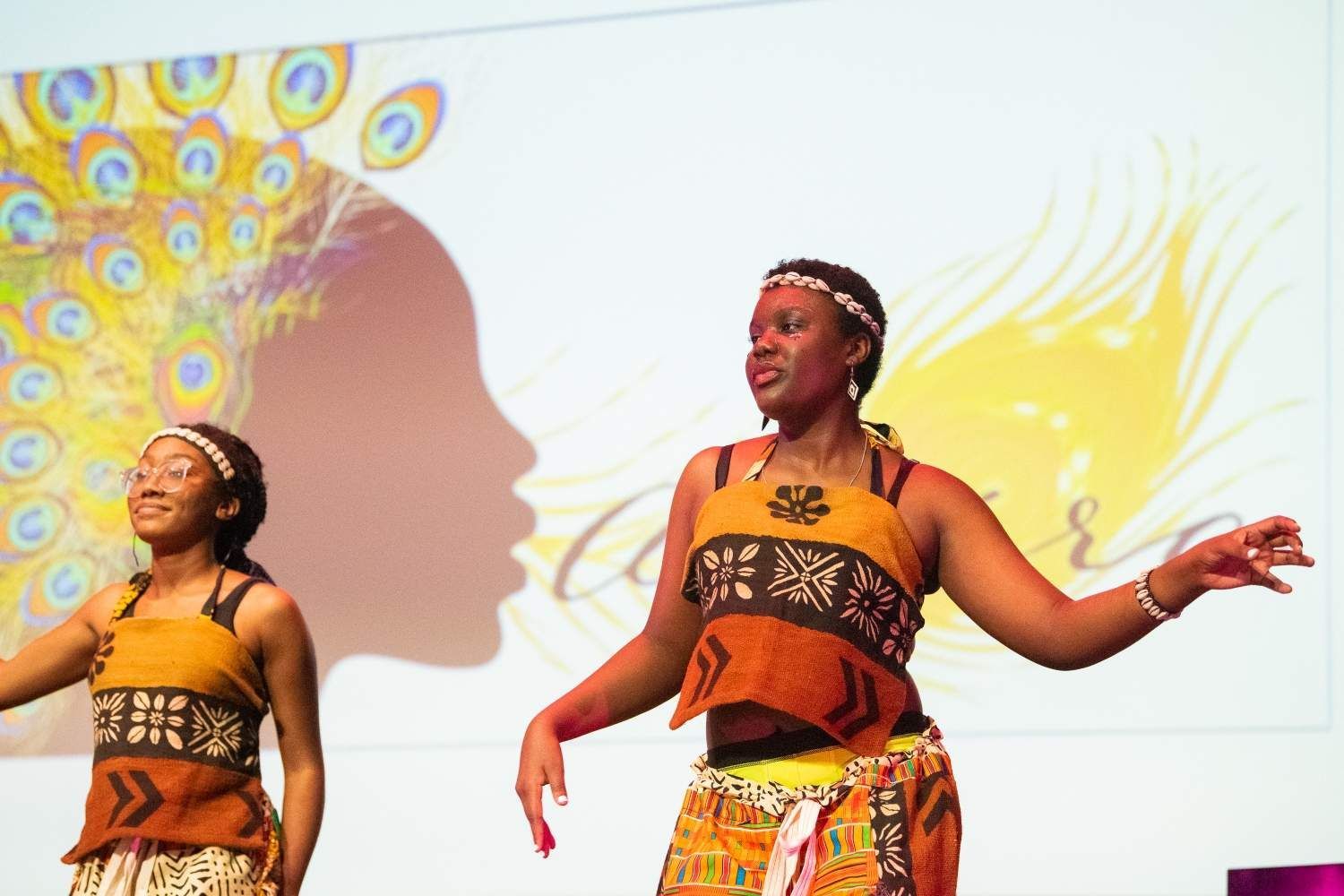 Two women in African attire performing on stage, backdrop features a silhouette and peacock design.